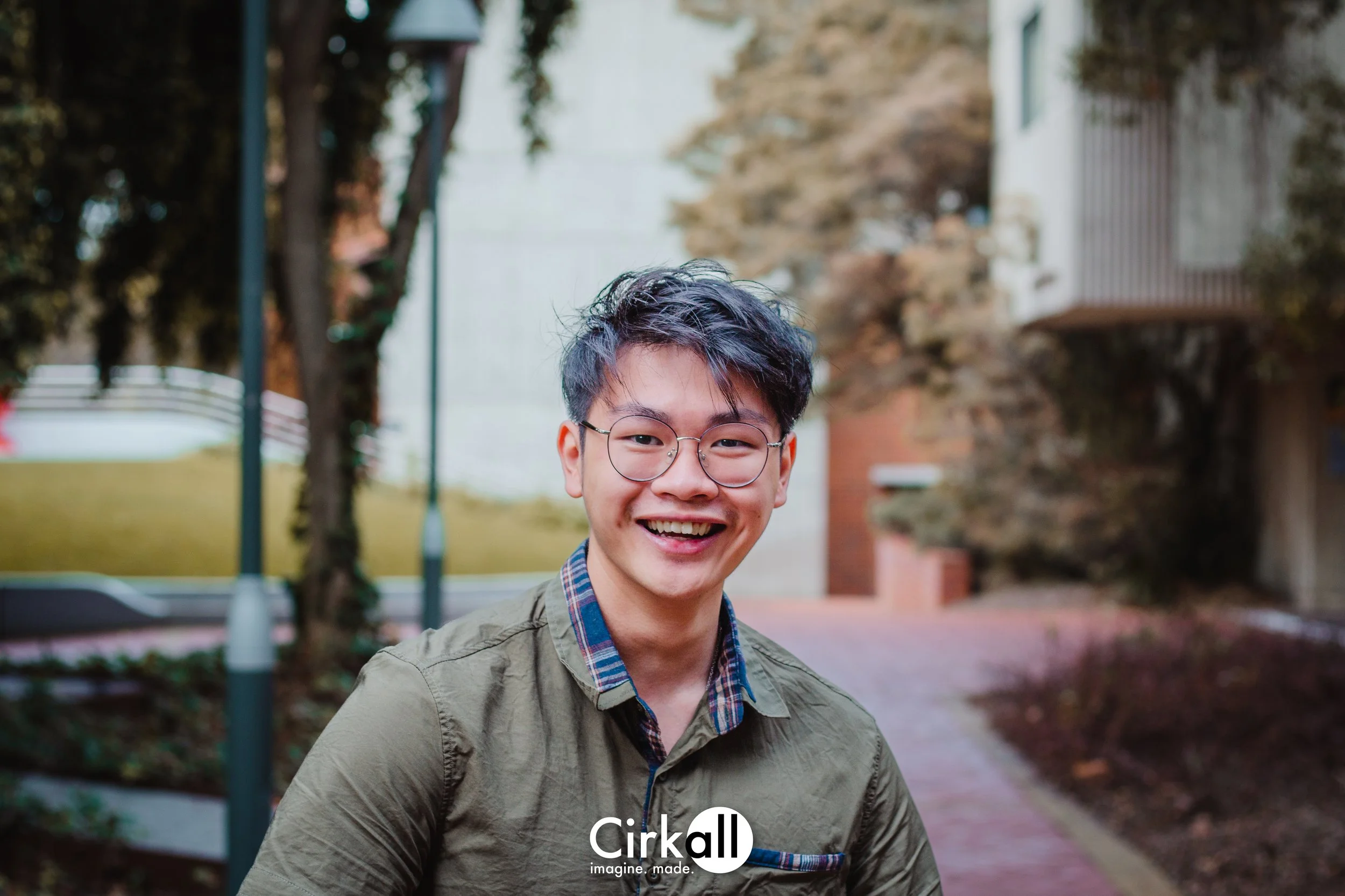 A young man with glasses smiling outdoors, with trees and a brick and white building in the background.