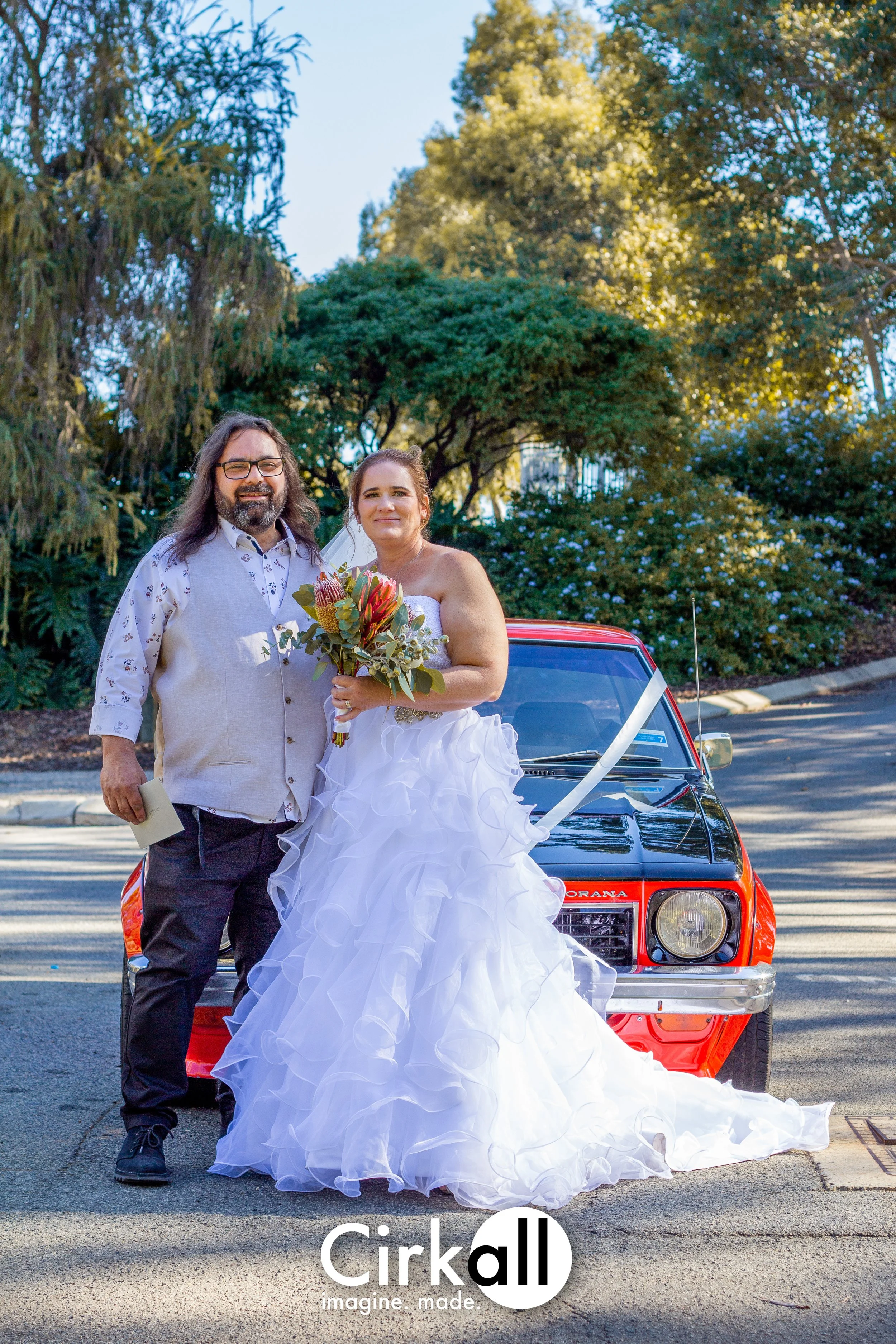 A newly married couple stands in front of a red vintage car outside, with lush green trees and a clear blue sky in the background. The bride is holding a bouquet of flowers and wearing a white wedding dress, while the groom is dressed in a light gray