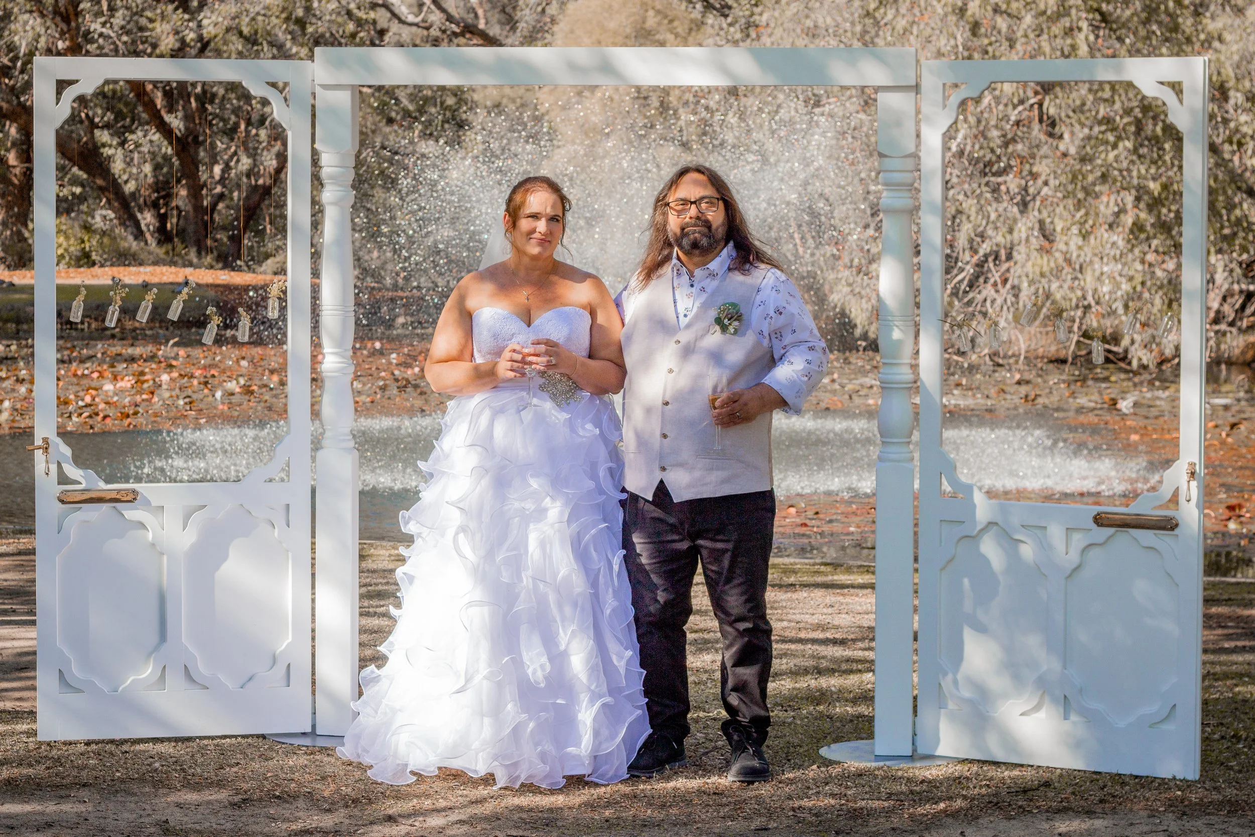 A bride in a white wedding gown and a man, possibly the groom, standing together outdoors under a decorative white archway with a water fountain in the background, people celebrating at a wedding.