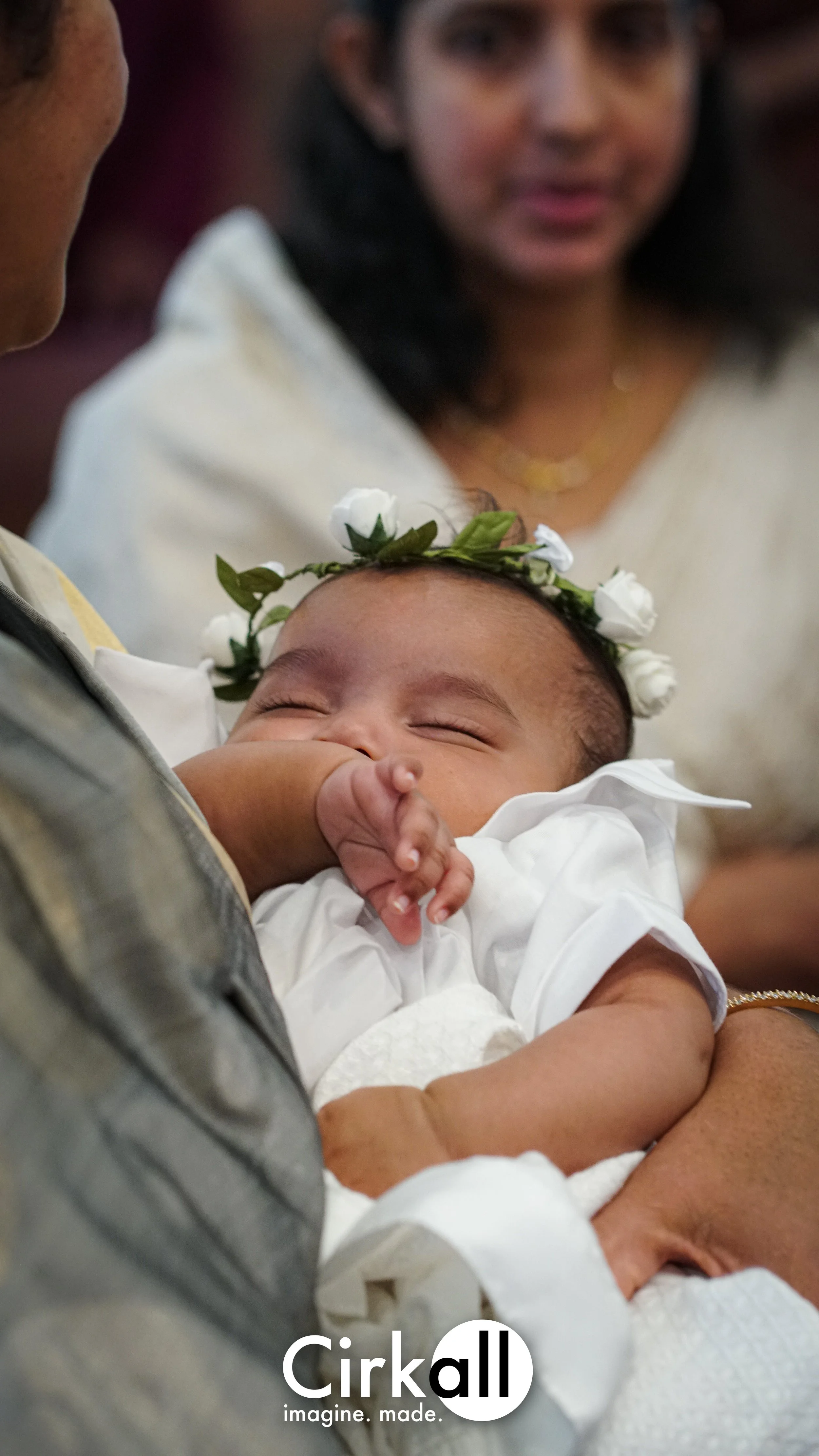 A sleeping baby wearing a white outfit and a floral headband being held by an adult. In the background, a woman with dark hair and a gold necklace looks on, with only part of her face visible.