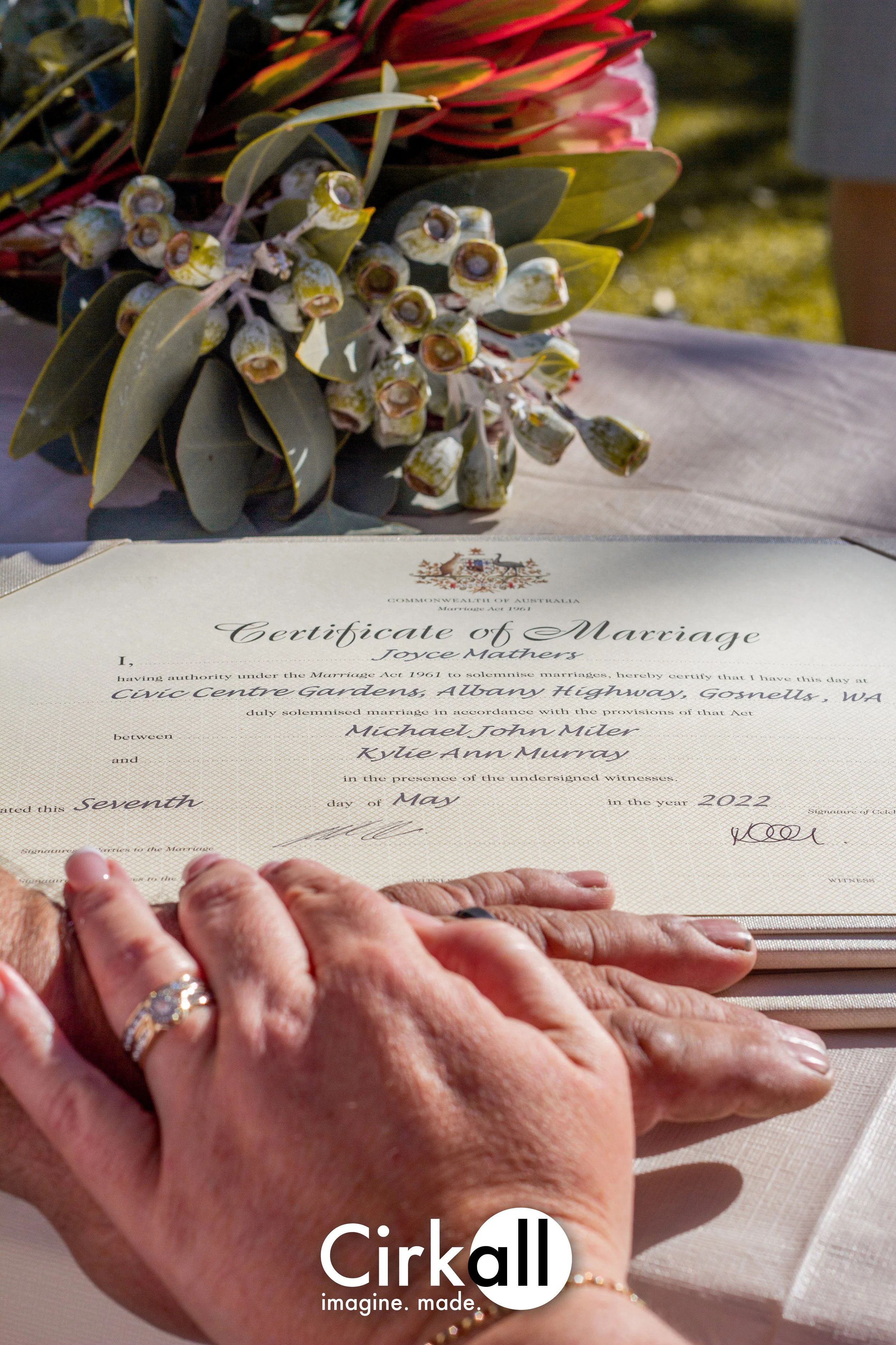 Close-up of a wedding certificate with two hands resting on it, one with a wedding ring, and a wedding bouquet with green leaves and pink flowers in the background.
