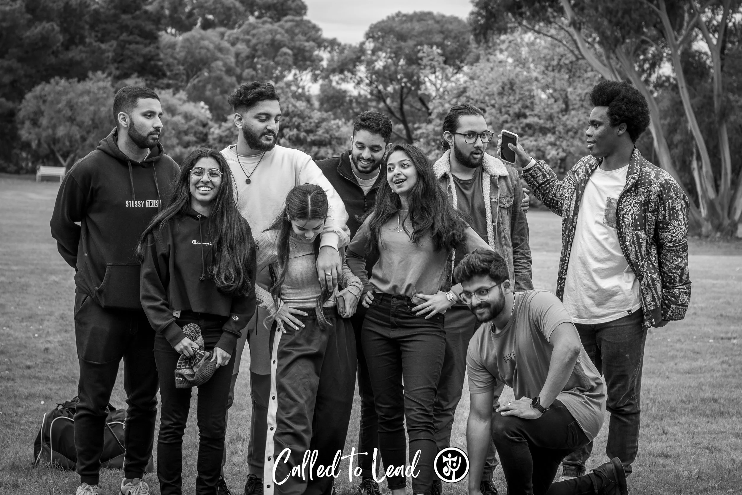 A group of nine young people taking a selfie together outdoors in a park. They appear happy and relaxed, with some smiling and others looking at the phone. The background features trees and a grassy area.