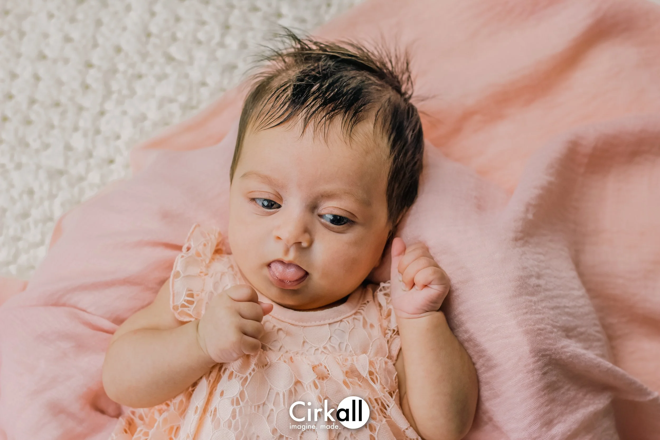 A baby girl lying on a soft pink blanket, wearing a lacy peach-colored dress, with her eyes open, tongue slightly sticking out, and fists clenched.