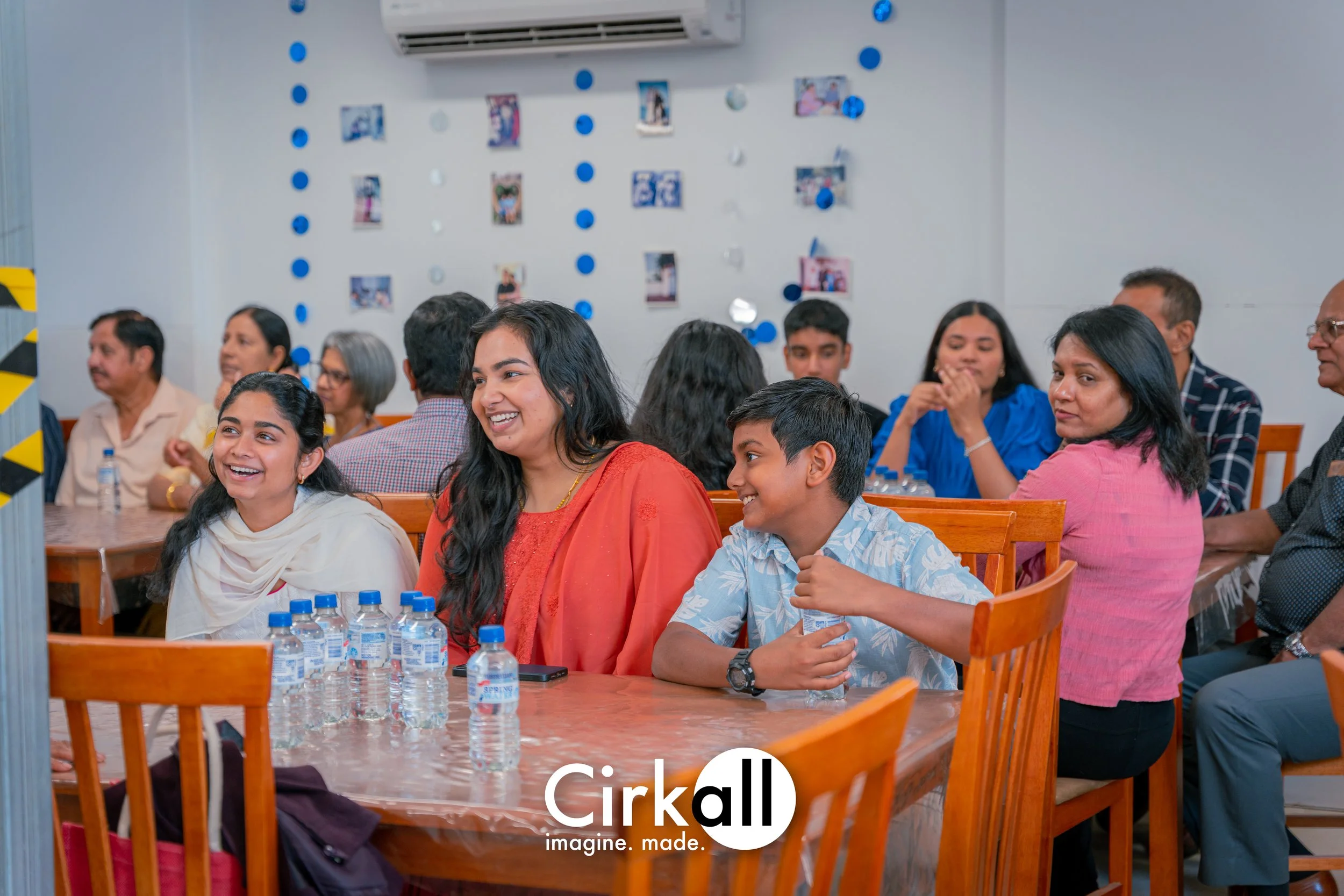Group of people sitting at a long wooden table, smiling and enjoying a gathering or event. Bottles of water are on the table, and a decorated wall with photos and blue and white ornaments is in the background.