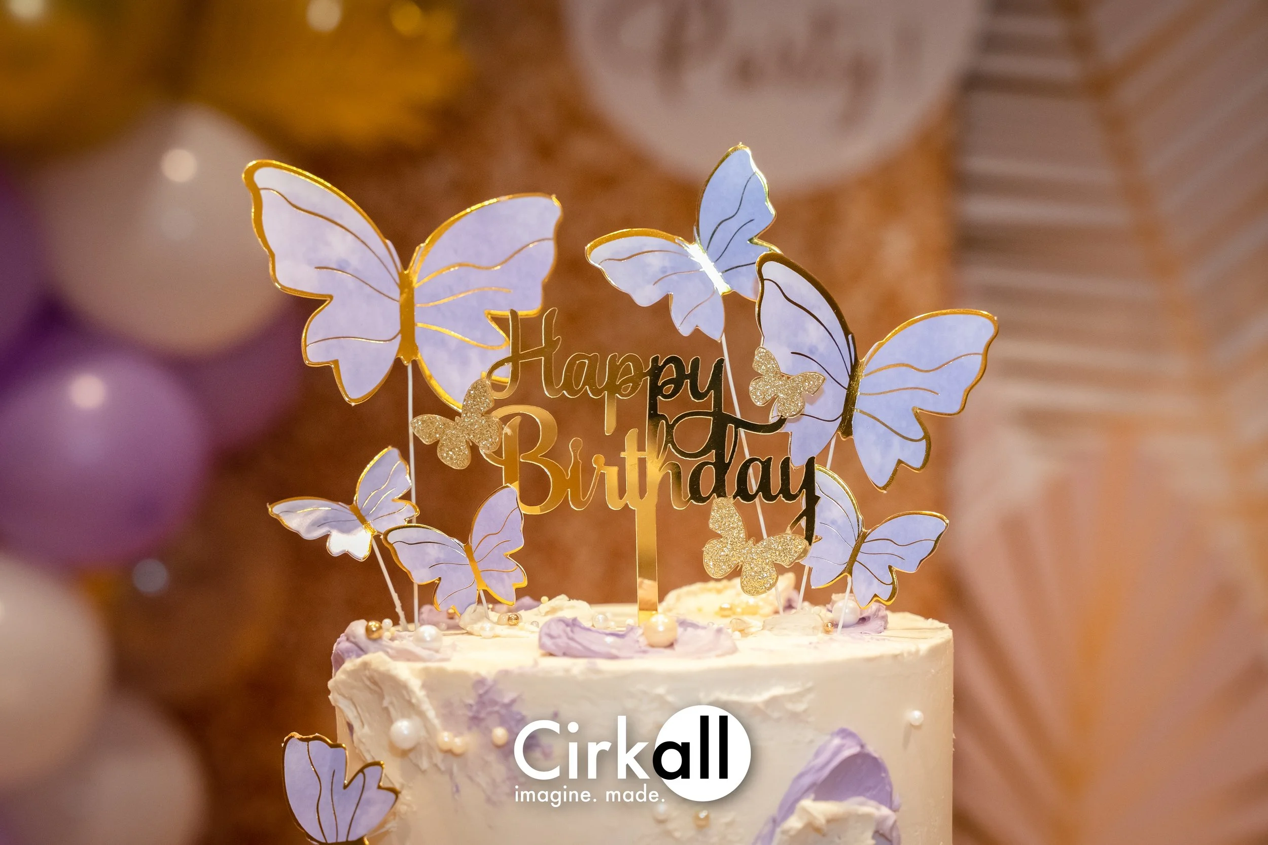 A birthday cake with a butterfly and flower cake topper that says "Happy Birthday," featuring purple butterfly and flower decorations on a white frosting cake.