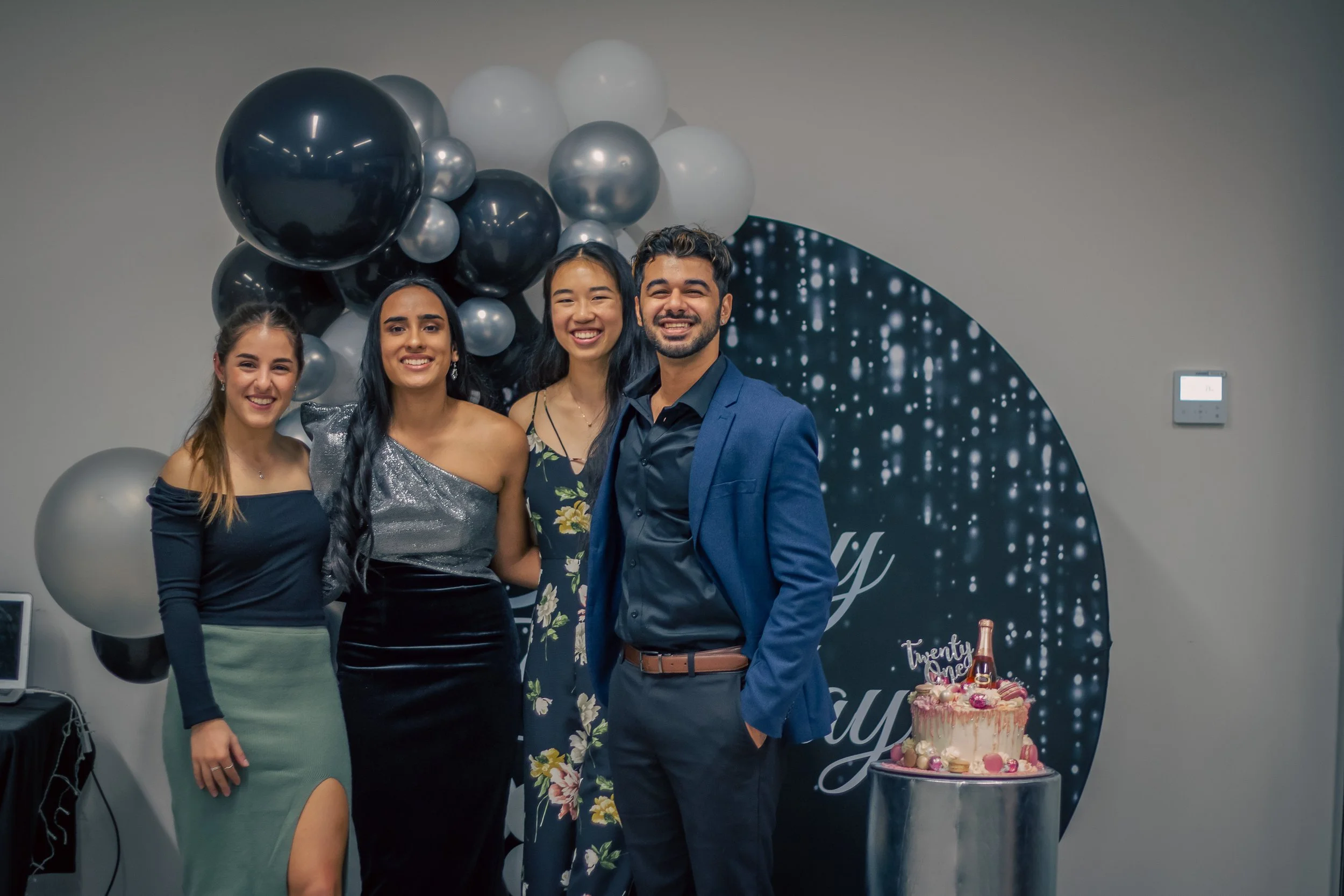 Four friends smiling at a celebration with black, white, and grey balloons in the background, a decorated cake on a stand, and a dark circular backdrop with lights and the word 'twenty' visible.