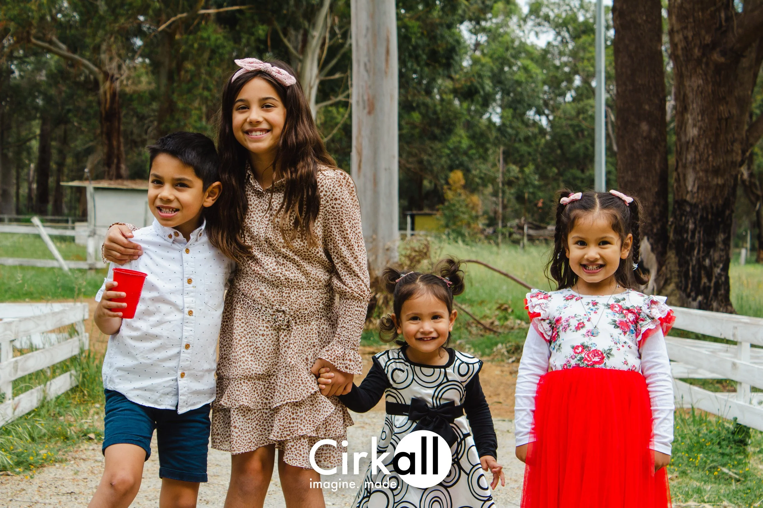 A woman and four young girls smiling outdoors in a park with trees and grass, holding hands and enjoying a sunny day.