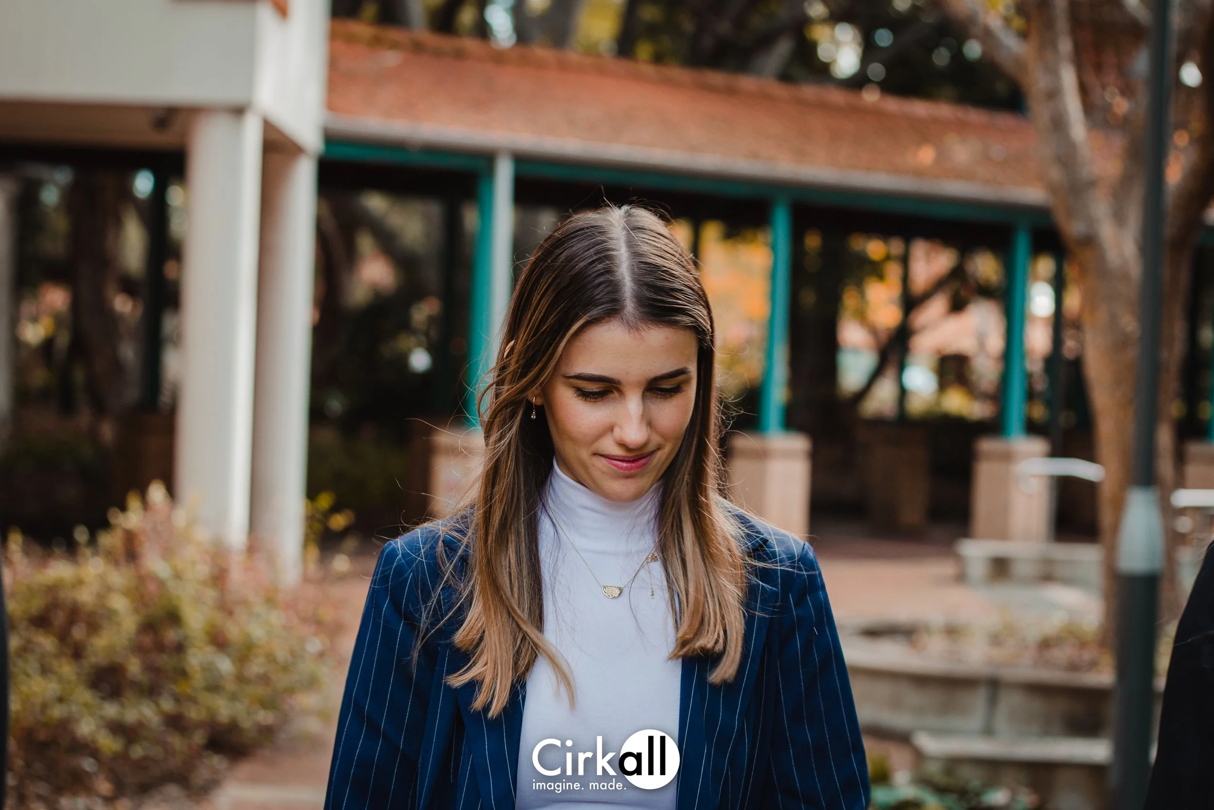 A young woman with long brown hair, wearing a navy pinstripe blazer and a white turtleneck, looking downward with a gentle smile in an outdoor park setting with trees, benches, and a structure with a red roof in the background.
