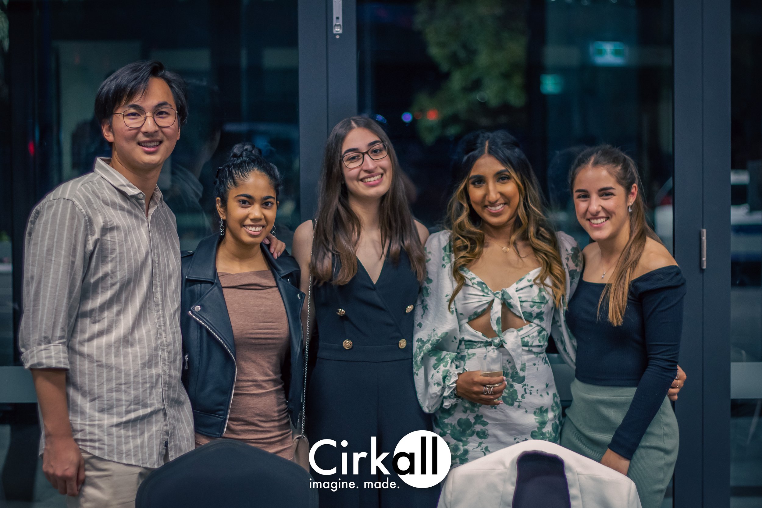 A diverse group of five young adults, three women and two men, smiling and posing indoors in front of glass doors during an evening event, with the Cirkall logo at the bottom.