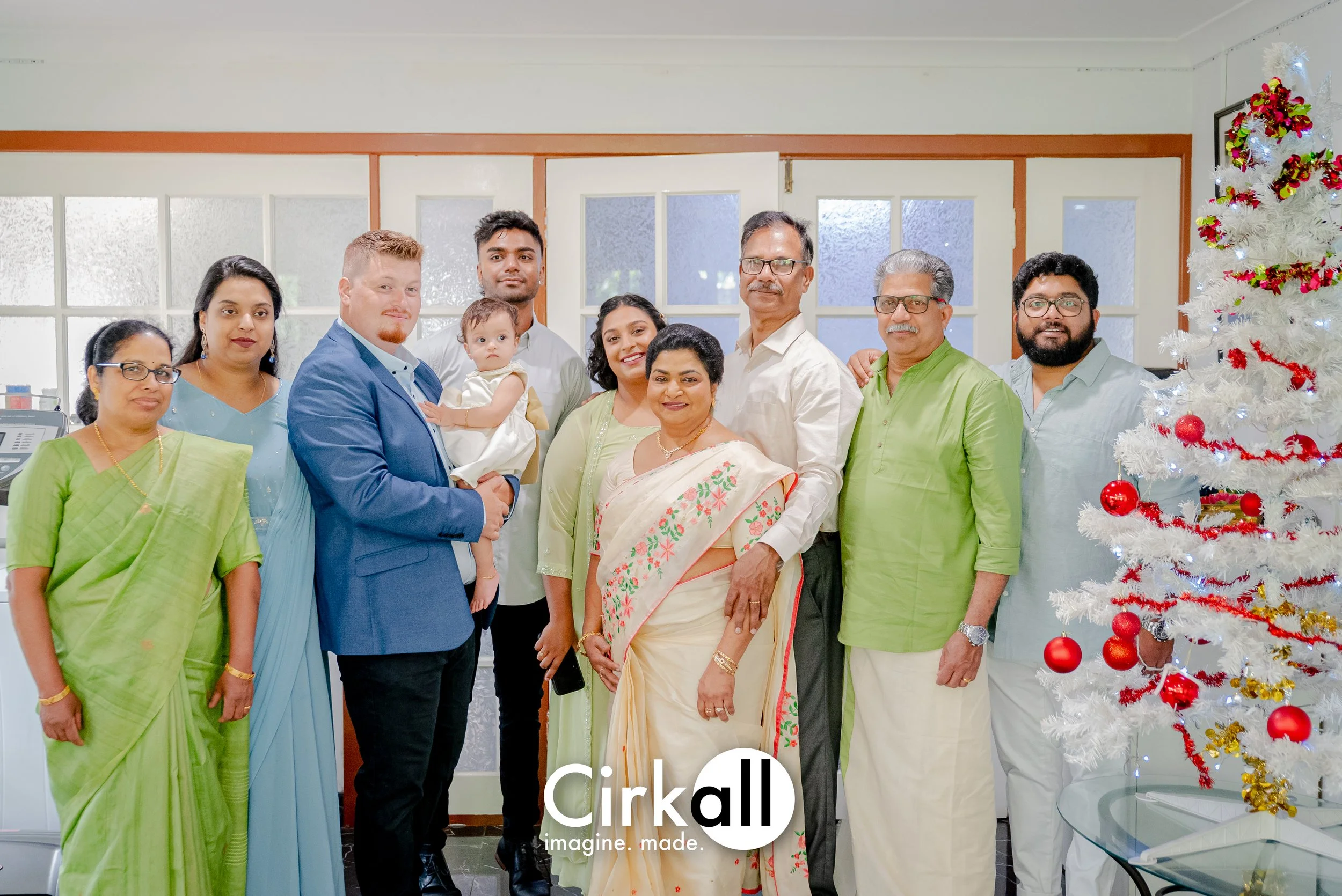 A multigenerational Indian family gathered indoors for a holiday celebration, with a decorated white Christmas tree visible on the right side of the image.