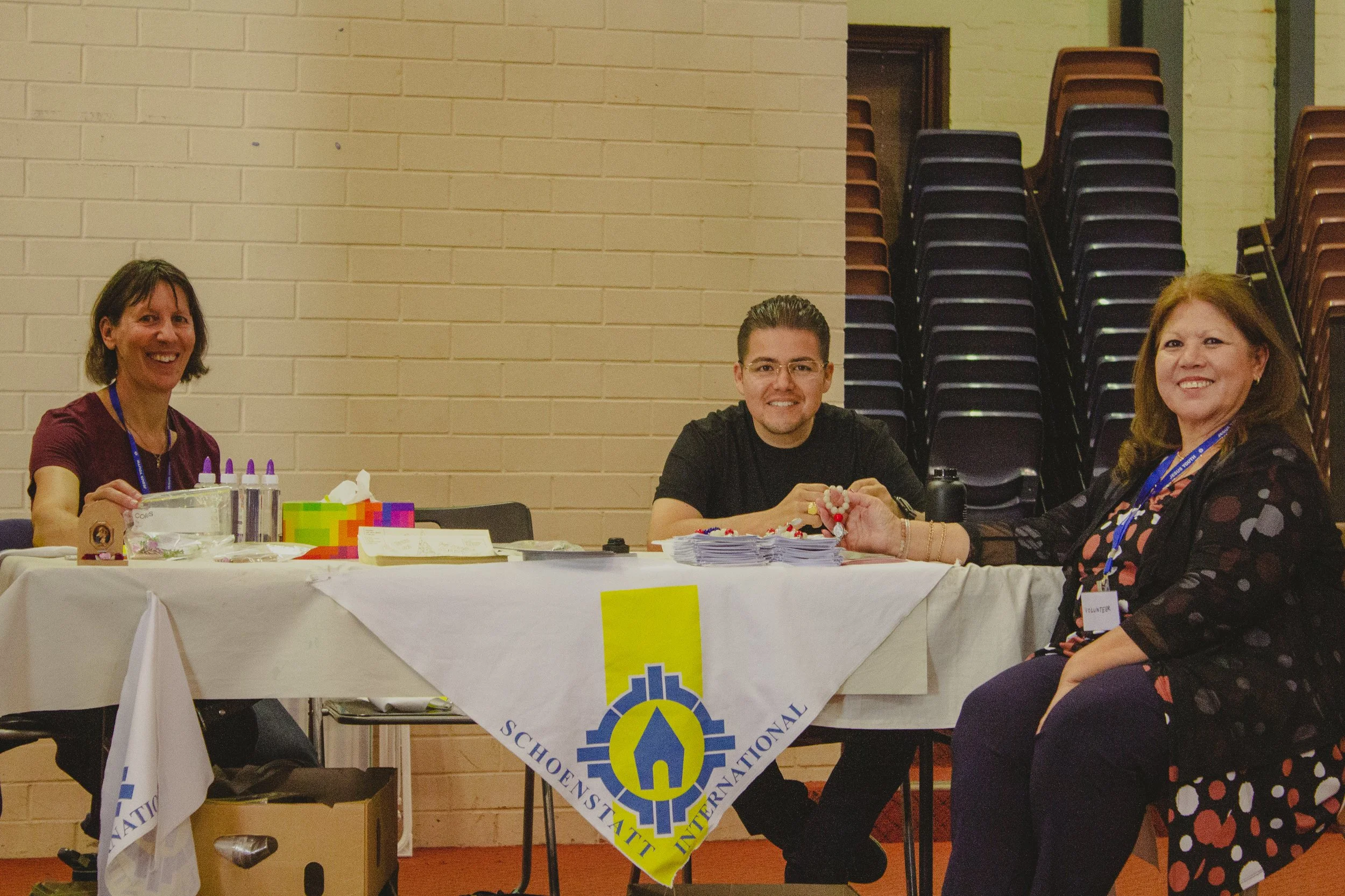 Three smiling volunteers sitting at a table with a white cloth, a Schoenstatt International banner, and various supplies, in an indoor setting with stacked chairs in the background.