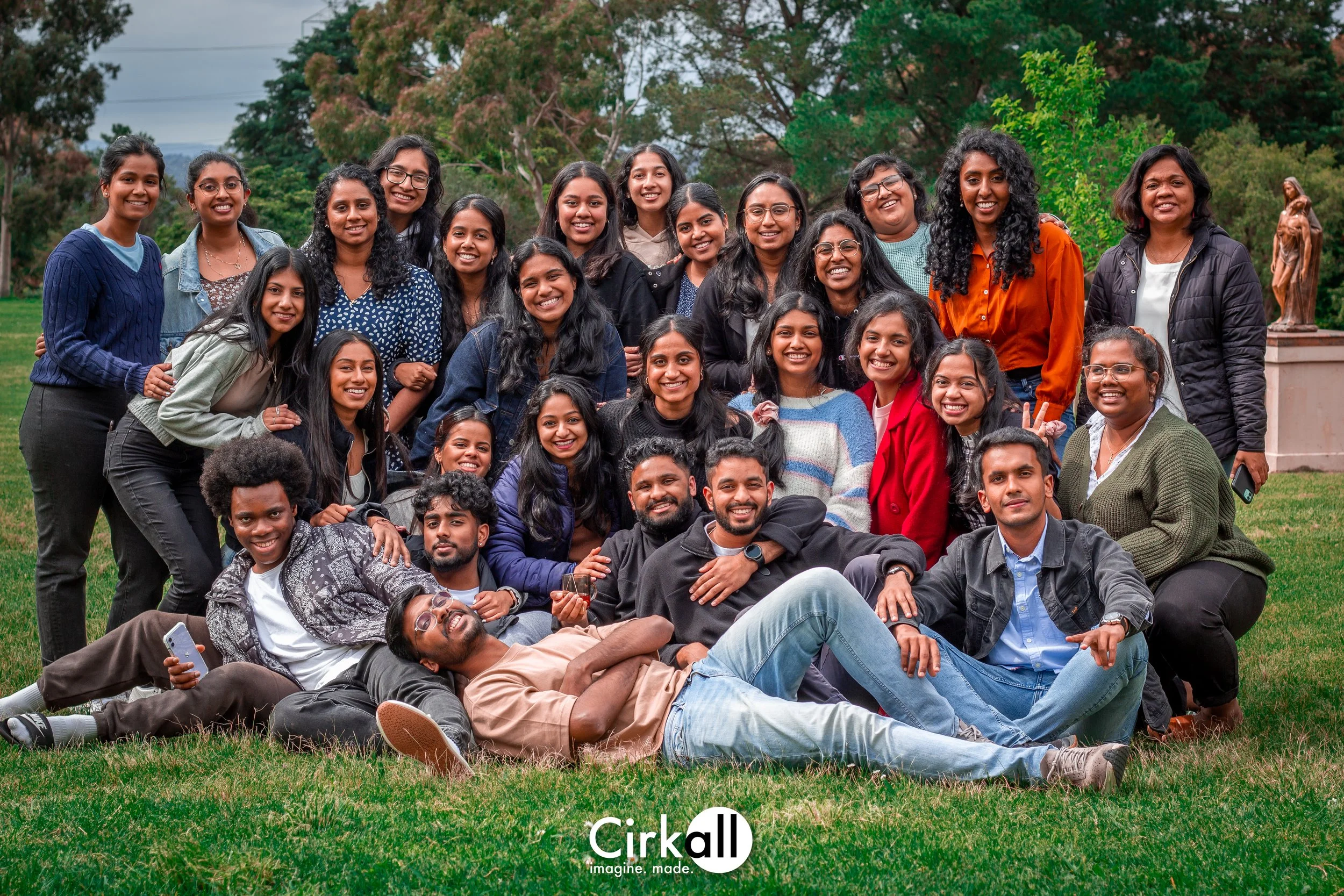 A group of diverse smiling young adults posing outdoors on grass, with trees and a statue in the background.