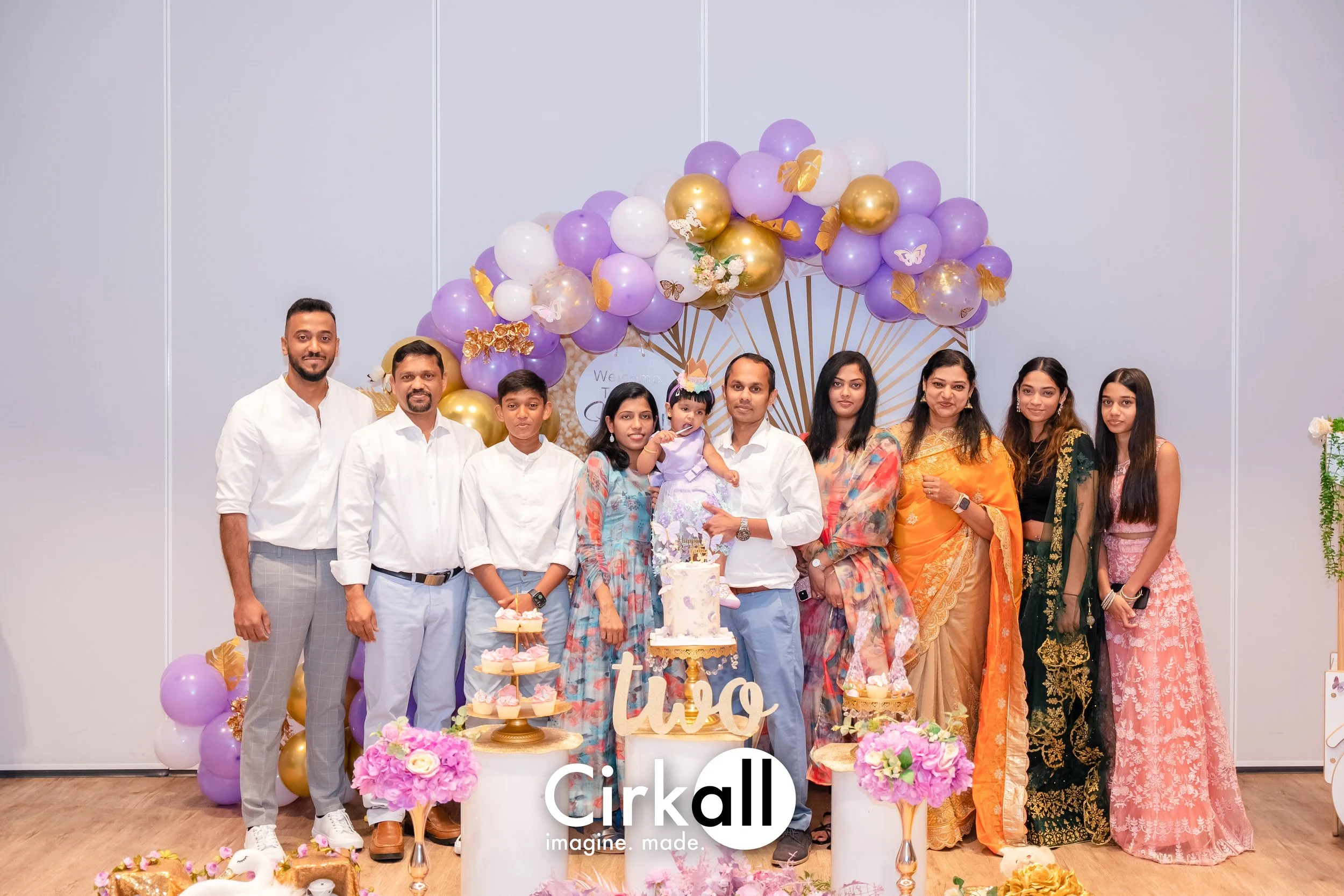 A group of ten people celebrating a child's second birthday, standing in front of a balloon arch with purple, white, and gold balloons, decorative butterflies, and flowers. The child is in the center, wearing a purple dress and a birthday crown, hold
