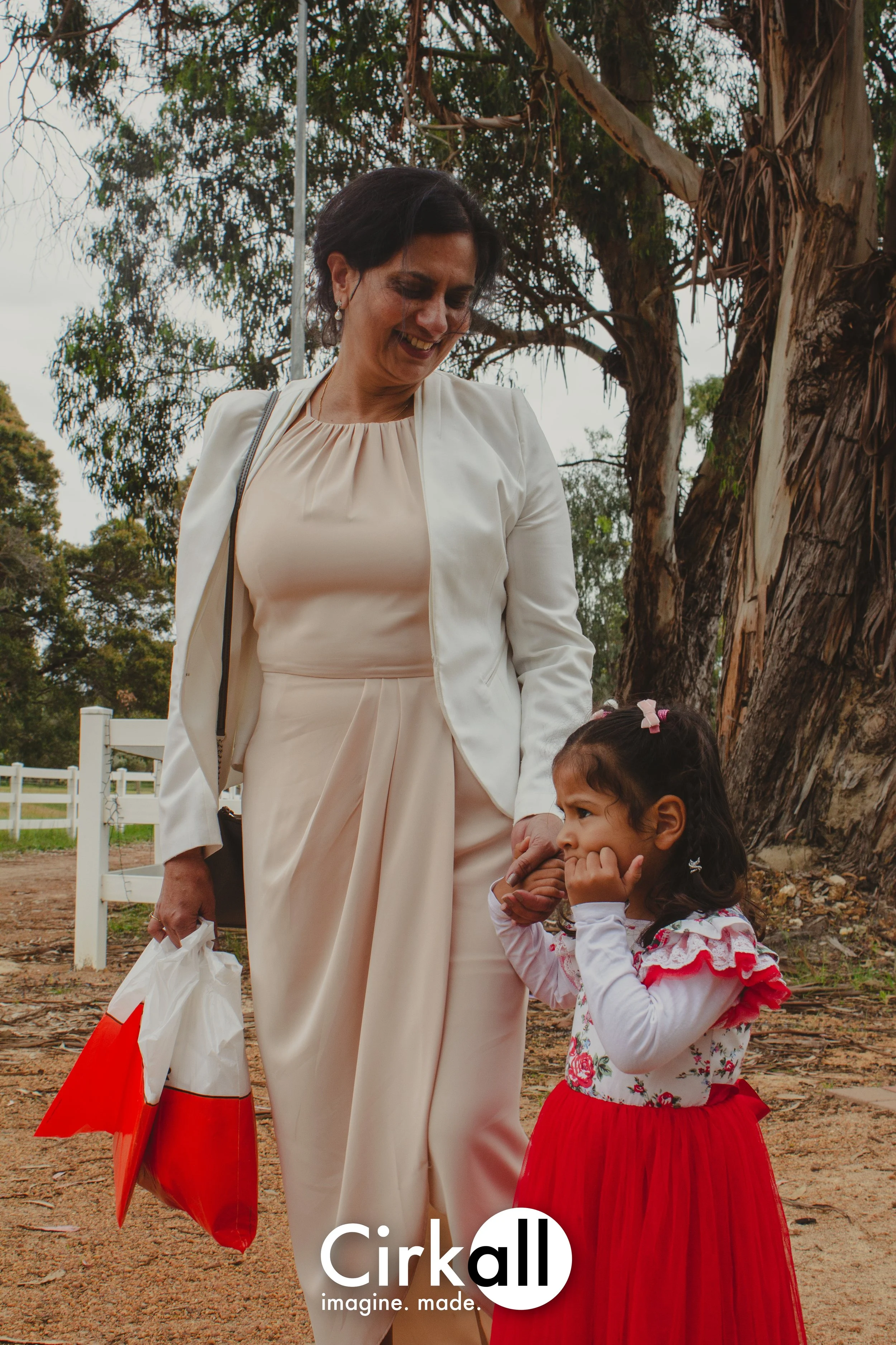 A woman smiling and holding a young girl’s hand outdoors near a large tree. The woman is wearing a cream-colored suit and the girl is dressed in a red and white dress with pink hair clips.