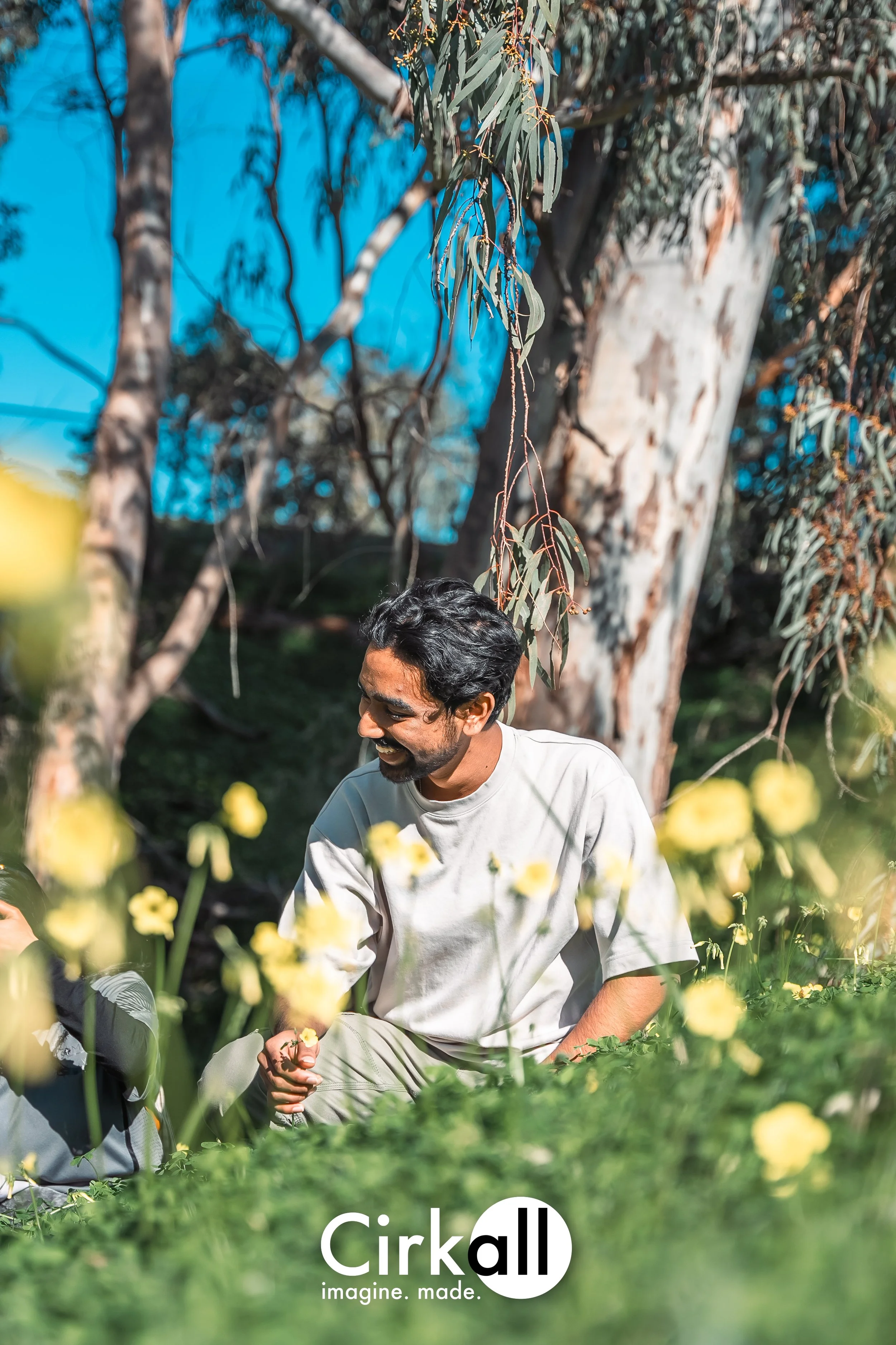 A man smiling and interacting with someone outdoors in a park or garden, surrounded by flowers and tall trees.
