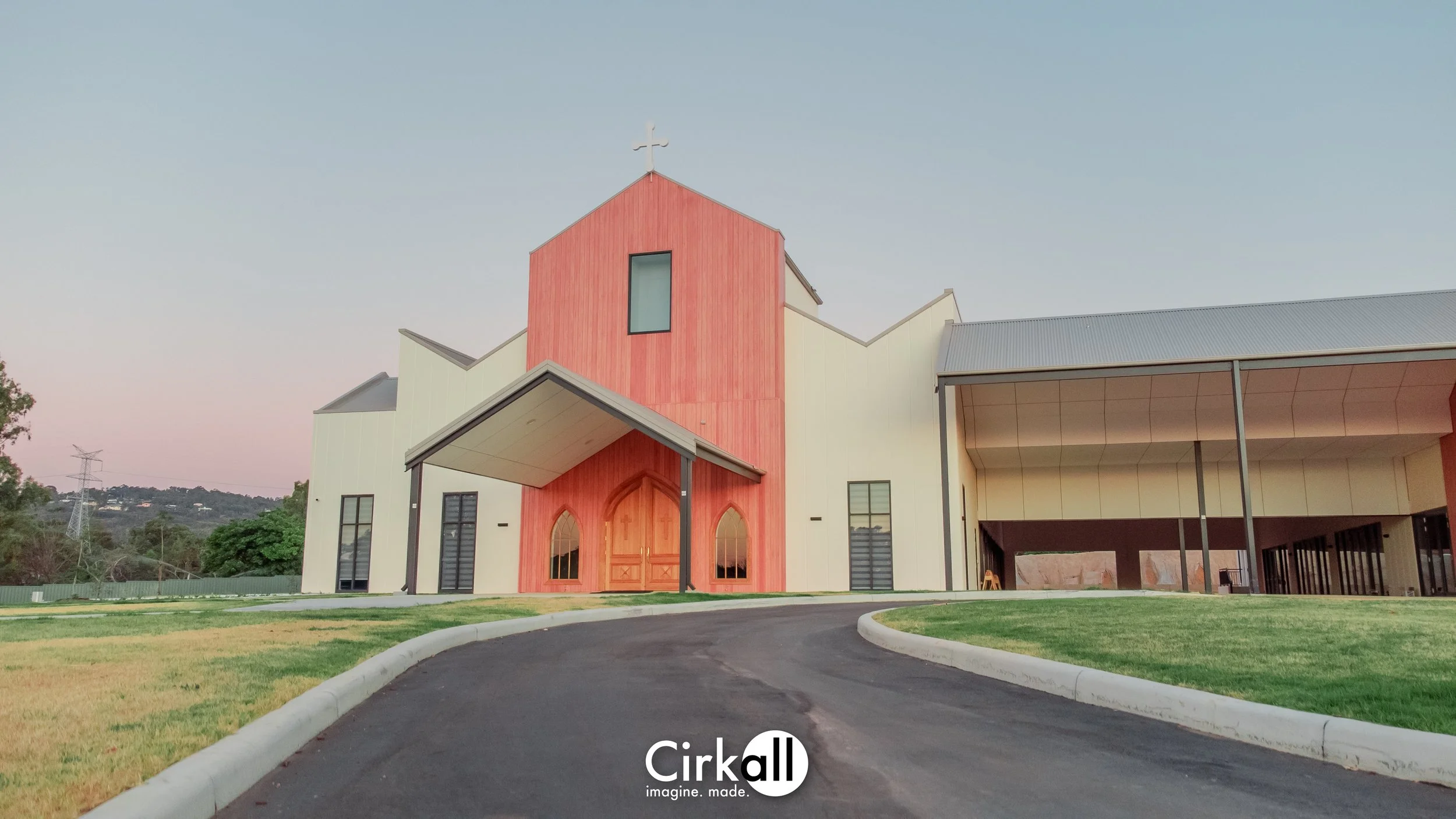 A modern church building with a red wooden front facade, arched windows and doors, and a cross on top. The church is surrounded by a curved driveway and green lawns, with a backdrop of trees and hills under a clear sky.