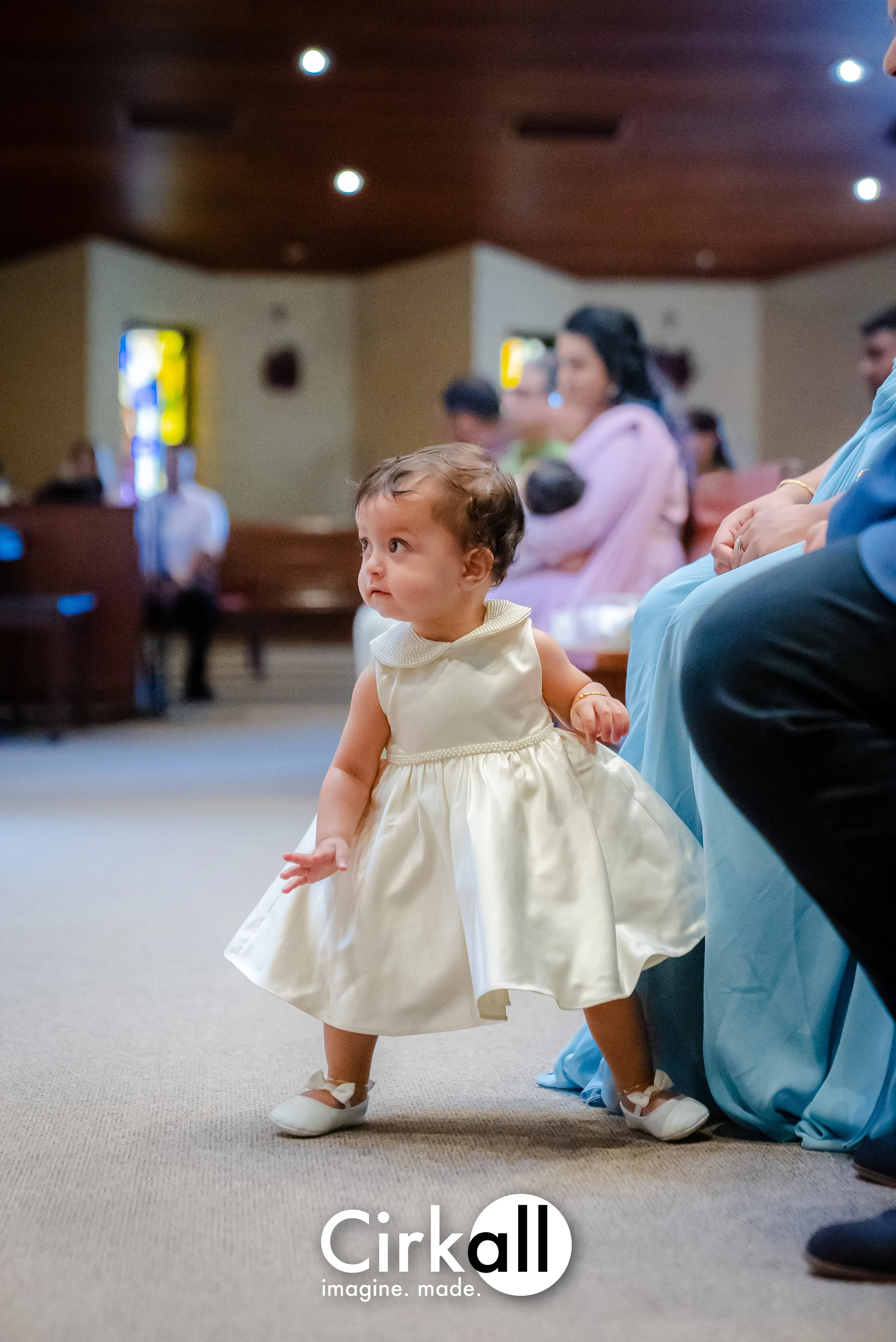A young girl in a white satin dress with a full skirt, white shoes, and a gold bracelet, walking in a church during a ceremony, with seated adults and stained glass windows in the background.