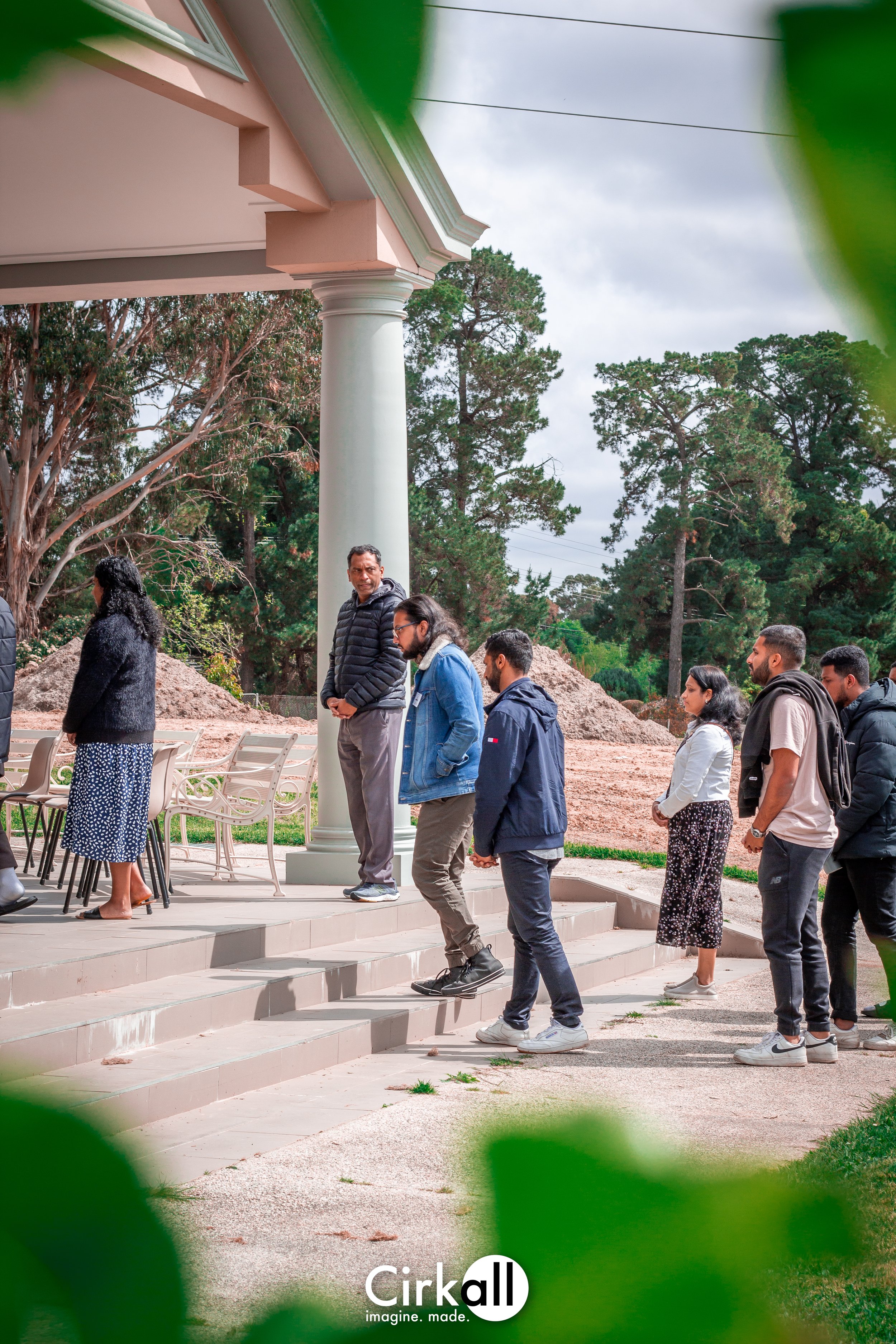 A group of people standing on the steps of a building with columns, outdoors, with trees in the background and a cloudy sky.