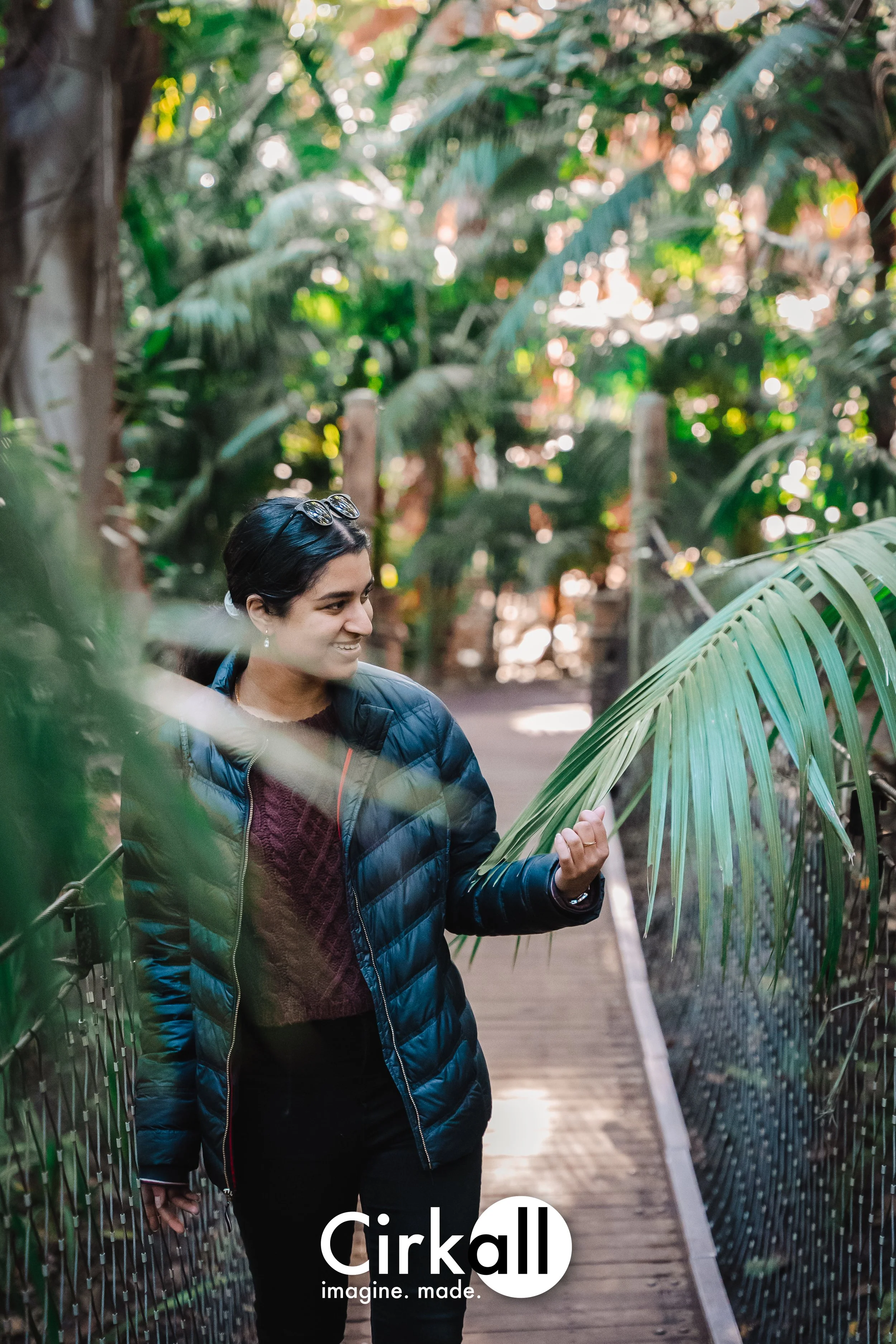 A woman in a blue jacket and burgundy sweater walking on a wooden pathway surrounded by lush green tropical plants, smiling and looking to the side.