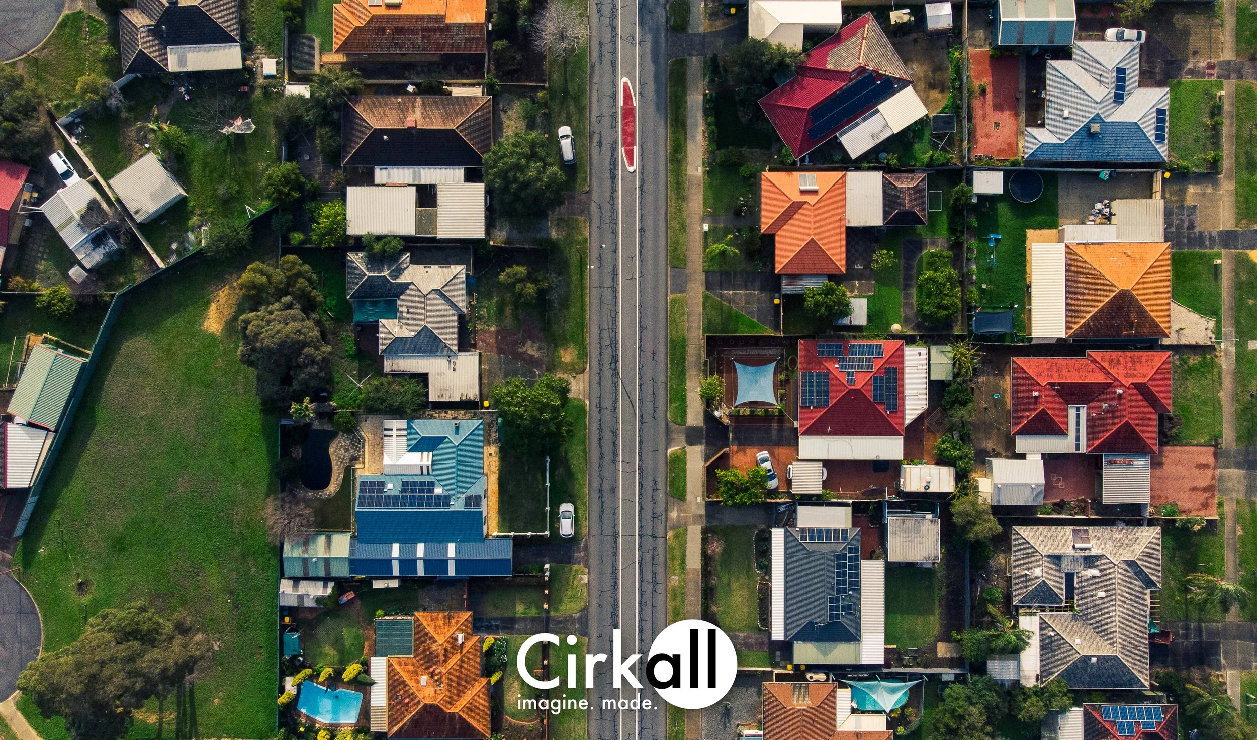 An aerial view of a suburban neighborhood with houses, trees, and yards, separated by a road with a tram or train track running down the middle.