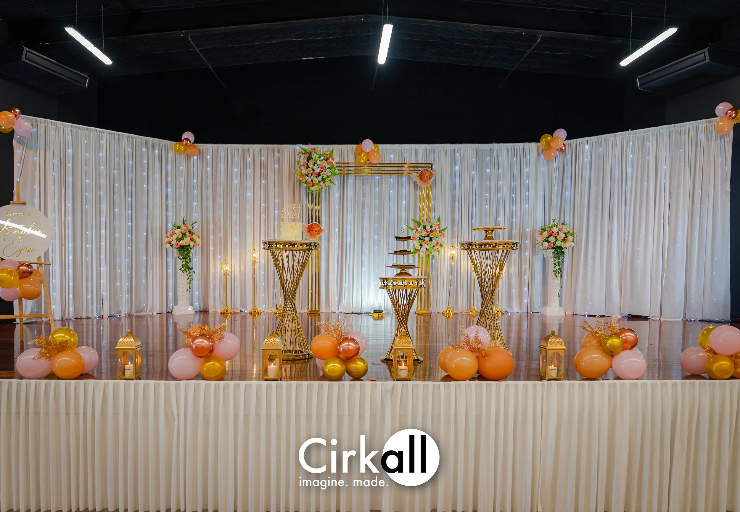 Decorated event stage with white curtains, string lights, pink and gold balloons, floral arrangements, lanterns, and gold tables, set for a celebration or wedding reception.