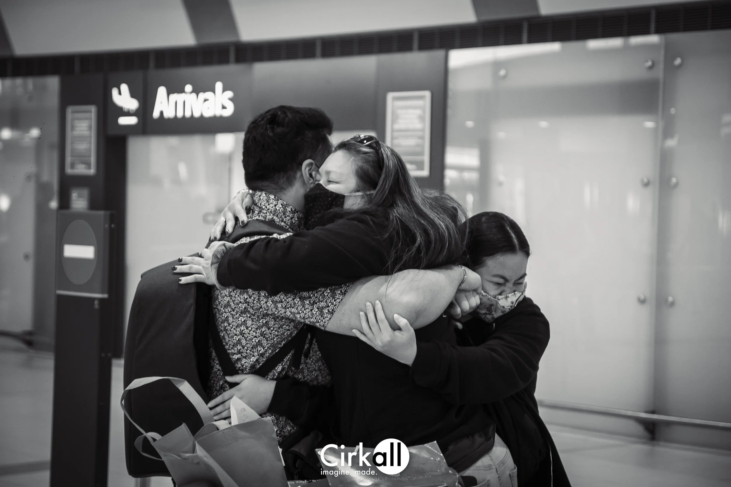 Three people embracing at an airport arrivals area, all wearing masks, with a sign that says 'Arrivals' in the background.