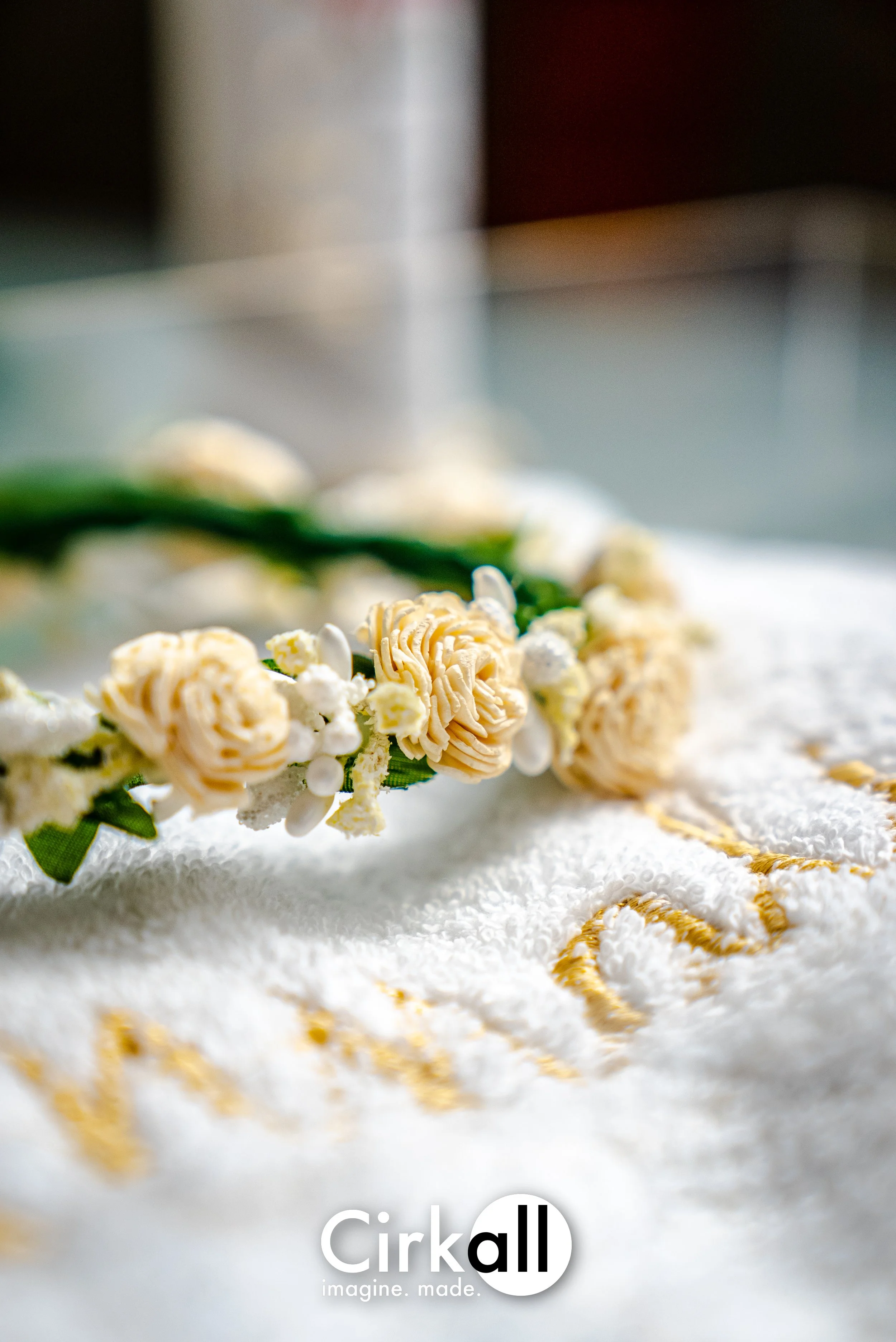 A white towel with gold embroidery and a flower crown with beige and white flowers resting on it.