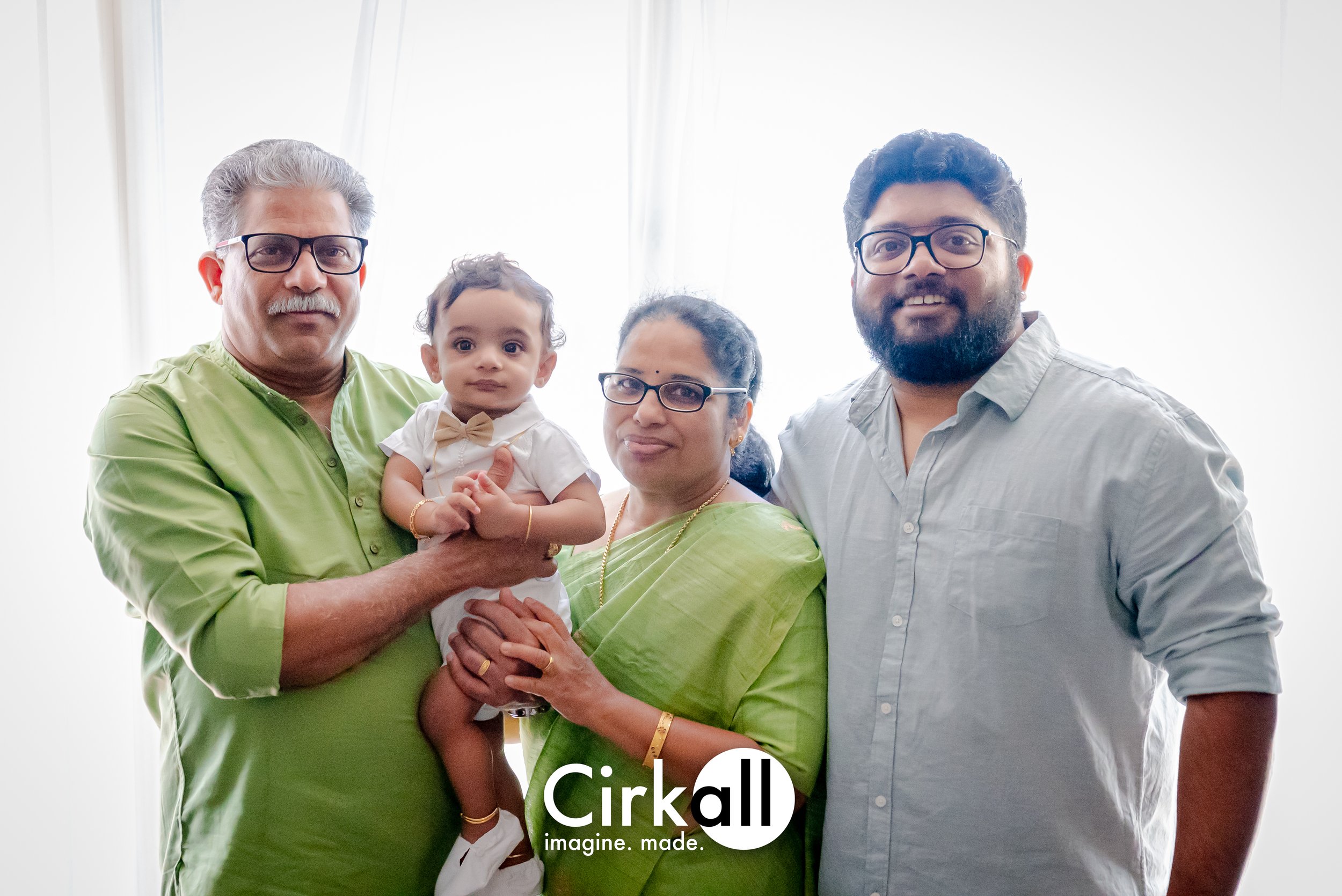 A family of four, including an elderly man, an elderly woman, a young man, and a toddler, all wearing glasses and dressed in light-colored clothing, standing together in front of a bright window.