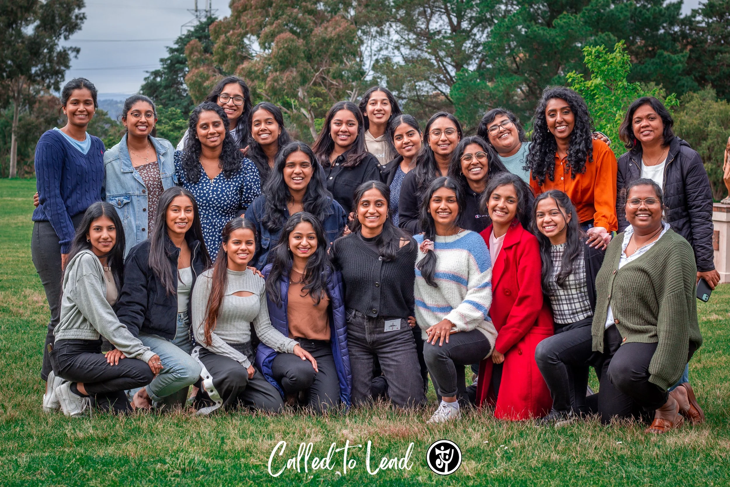 Group of young women outdoors on a grassy field, standing and kneeling, smiling at the camera. Trees and cloudy sky in the background. Text at the bottom reads 'Called to Lead' with a logo.