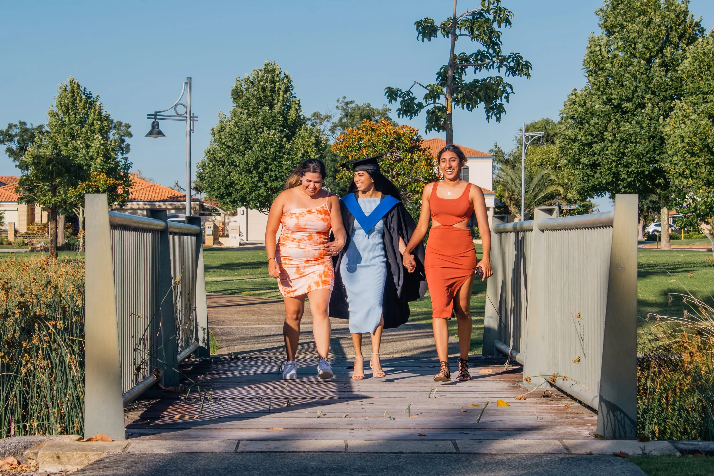 A woman in a graduation gown and cap walking with two women in summer dresses on a bridge in a park.