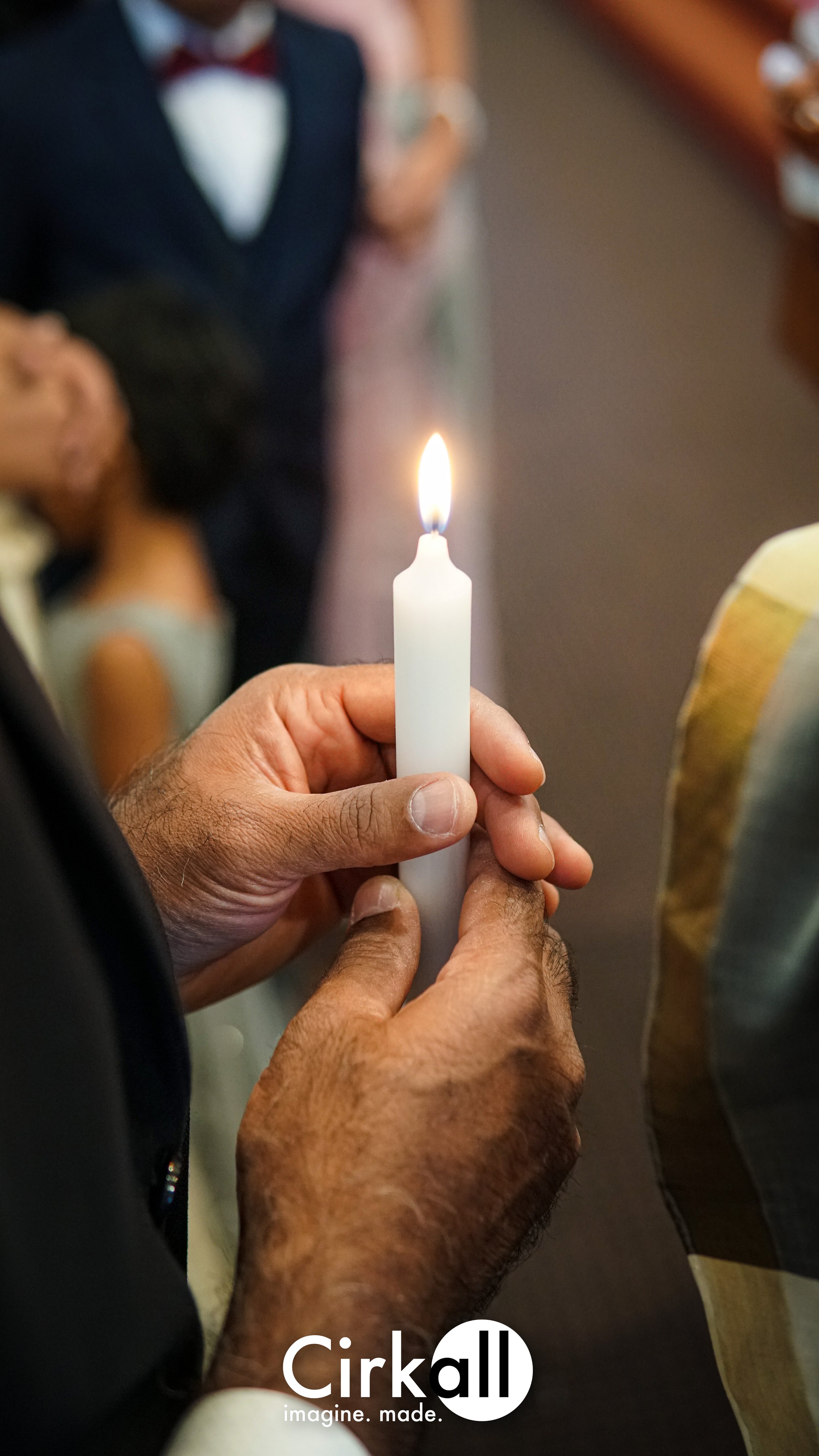 A person holding a white candle with a small flame, surrounded by blurred people in formal attire at what appears to be a ceremony or celebration.