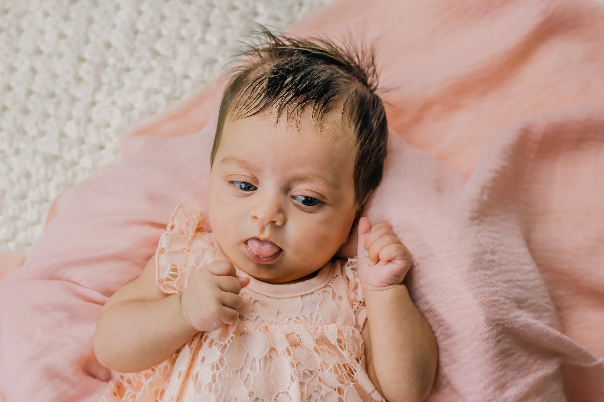 A baby lying on a pink blanket with a textured white surface nearby, wearing a peach-colored dress with lace details, sticking out tongue and looking to the side.