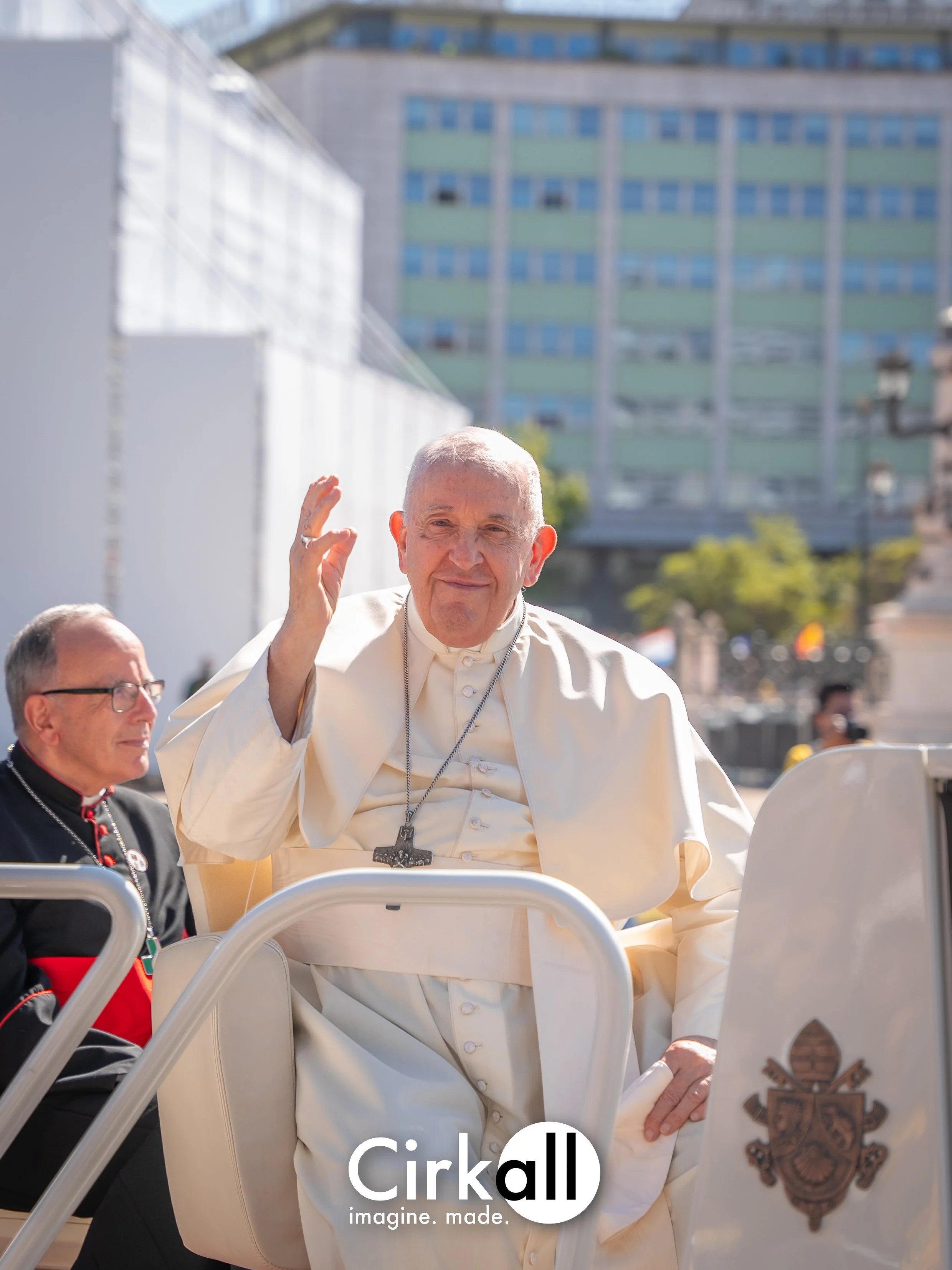 Pope Francis waving as he rides in a popemobile with a woman nearby, with urban buildings in the background.