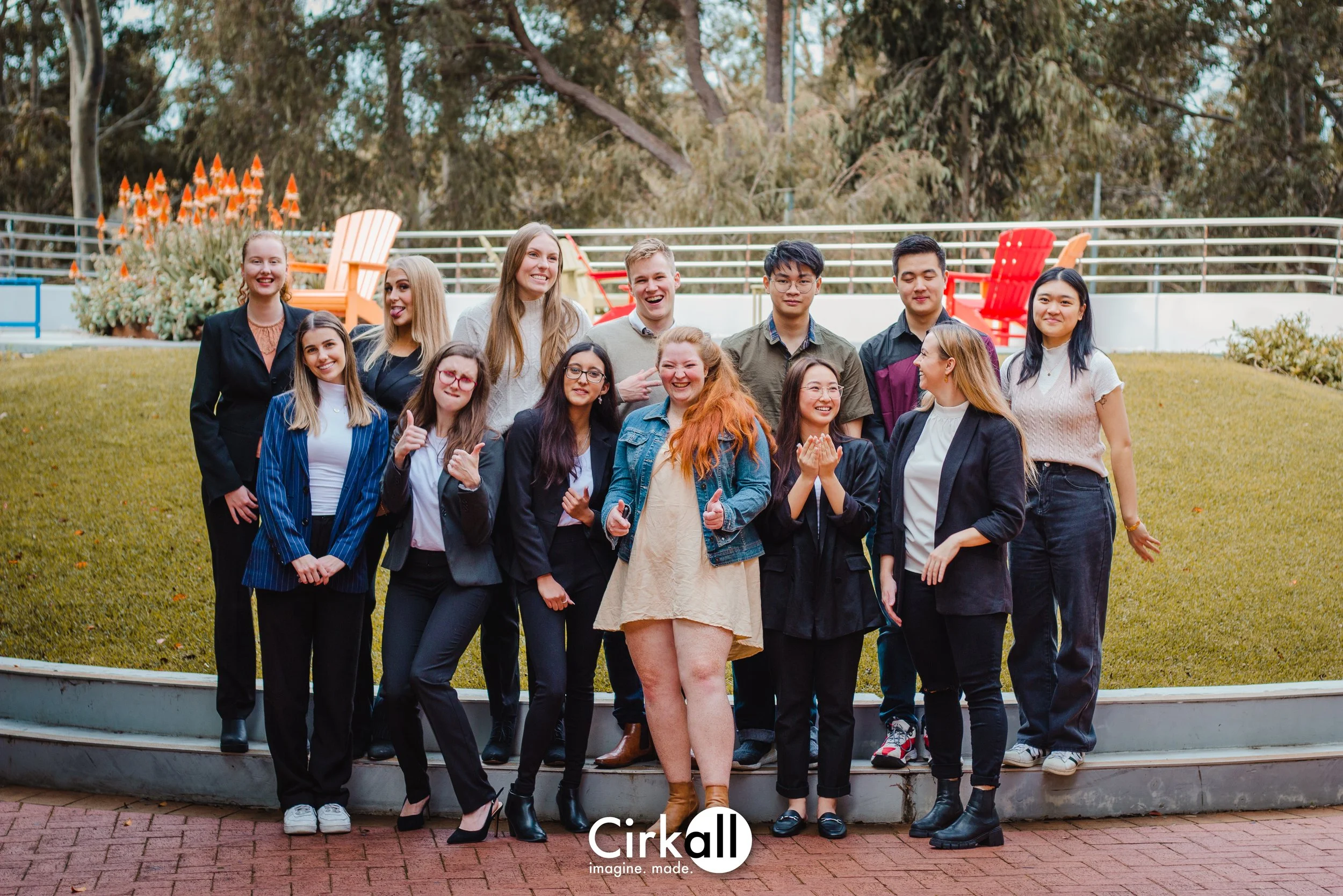 A group of diverse young adults pose together outdoors on a grassy area with trees in the background. They are smiling and dressed in a mix of casual and semi-formal clothing, standing on a curved concrete platform. Some are giving thumbs up or makin