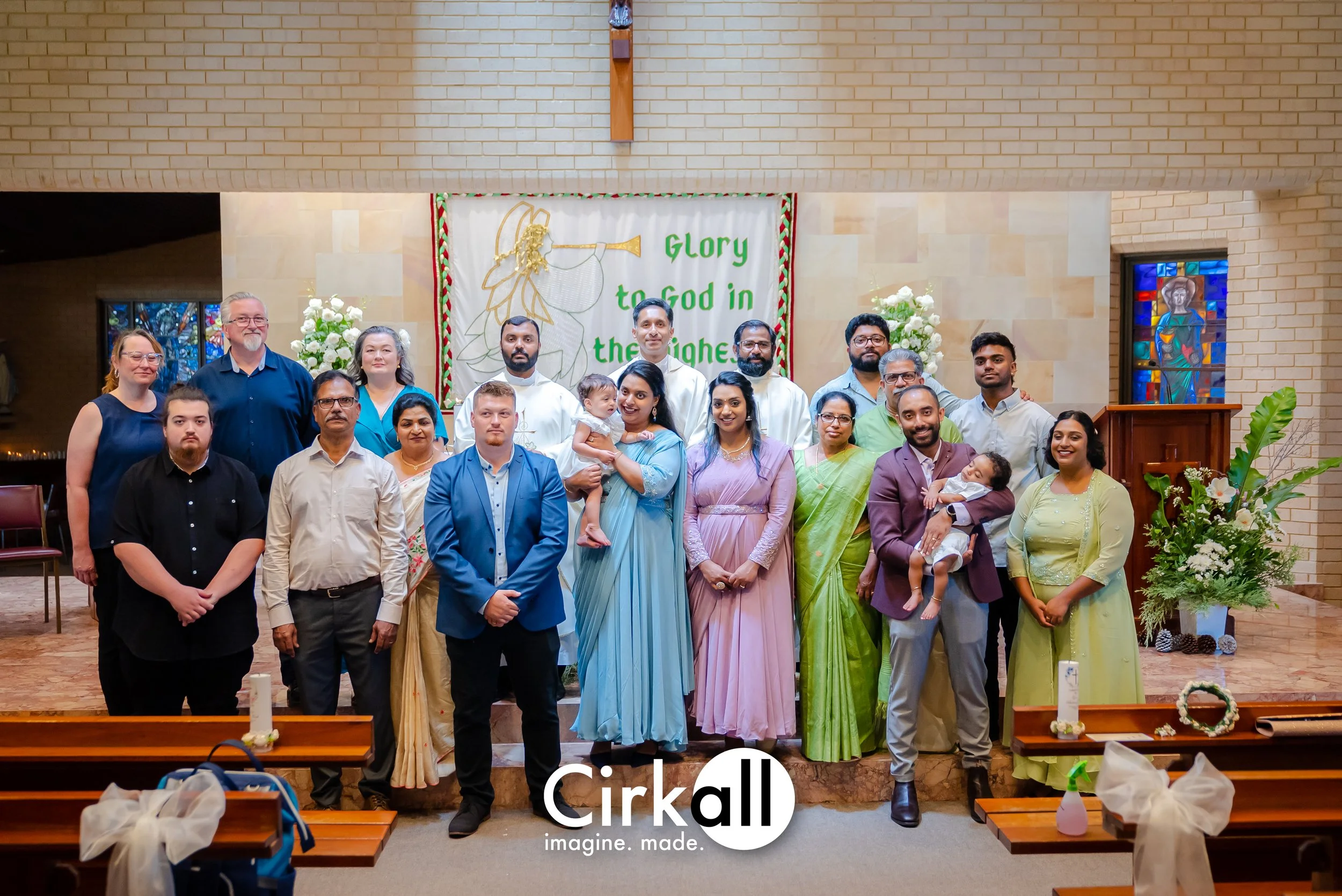 A group of people gathered inside a church, posing for a photo. The background features a banner with an angel drawing and the text "Glory to God in the highest." Some people are wearing traditional Indian attire, and there are floral arrangements ar