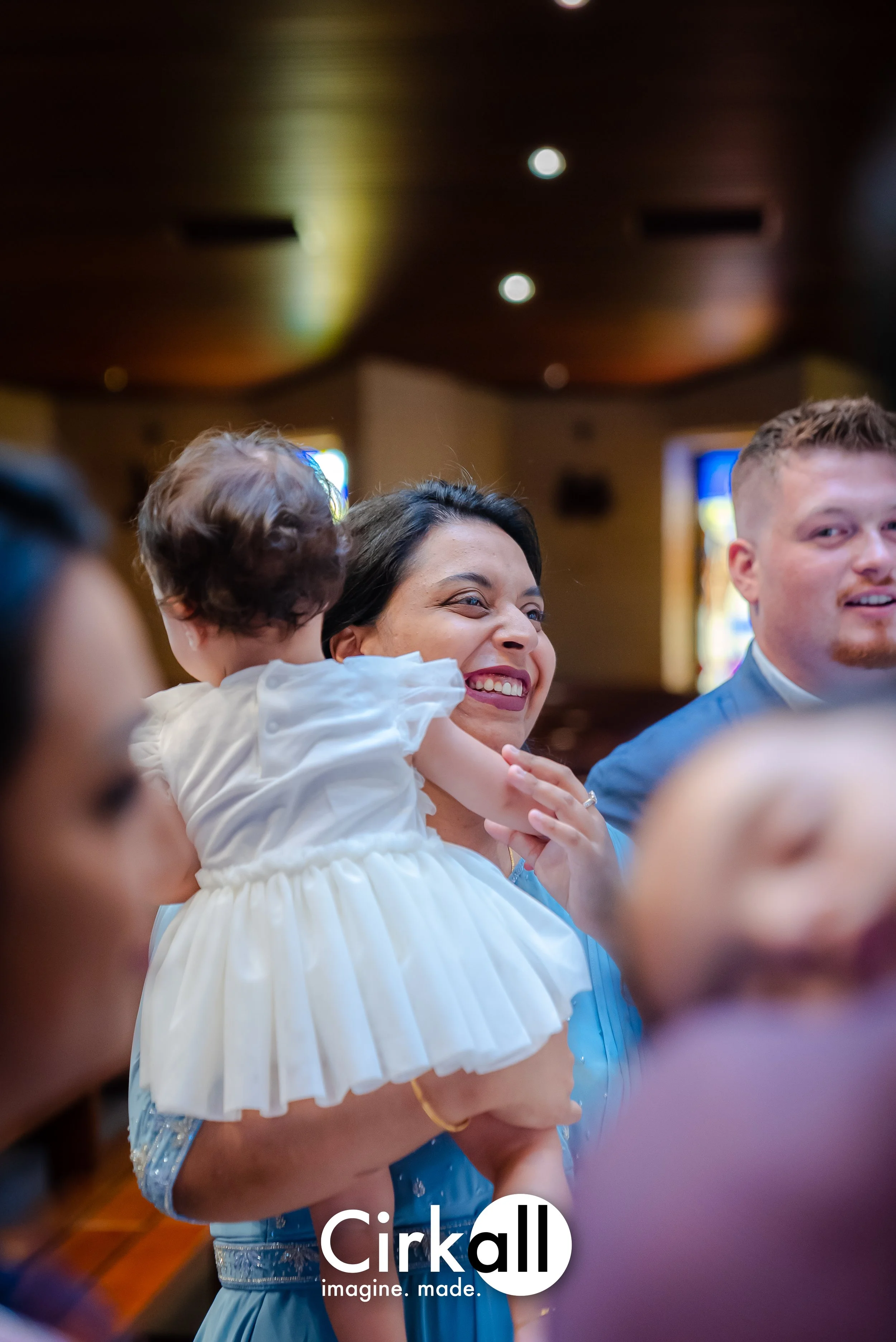 A woman holding a young child in a white dress, smiling during a celebration in a dimly-lit indoor setting.