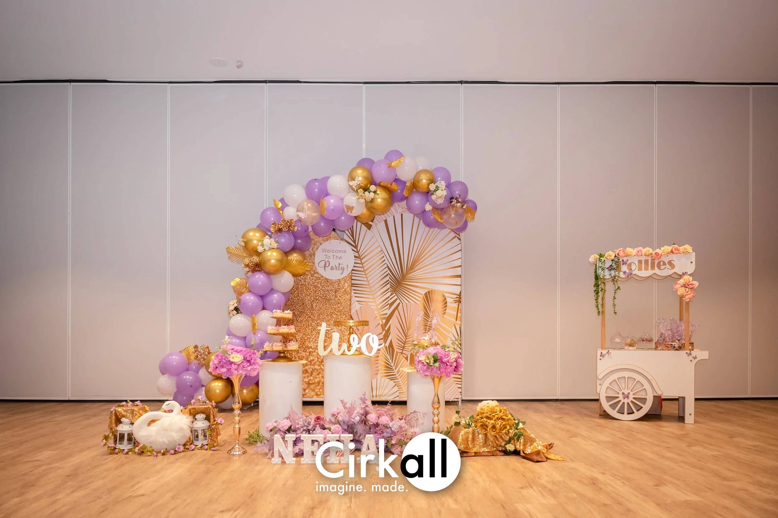 Decorations for a celebration with a purple, white, and gold balloon arch, pink flowers, a swan figure, and signs that read 'two' and 'NEO,' with a backdrop featuring tropical leaf patterns.