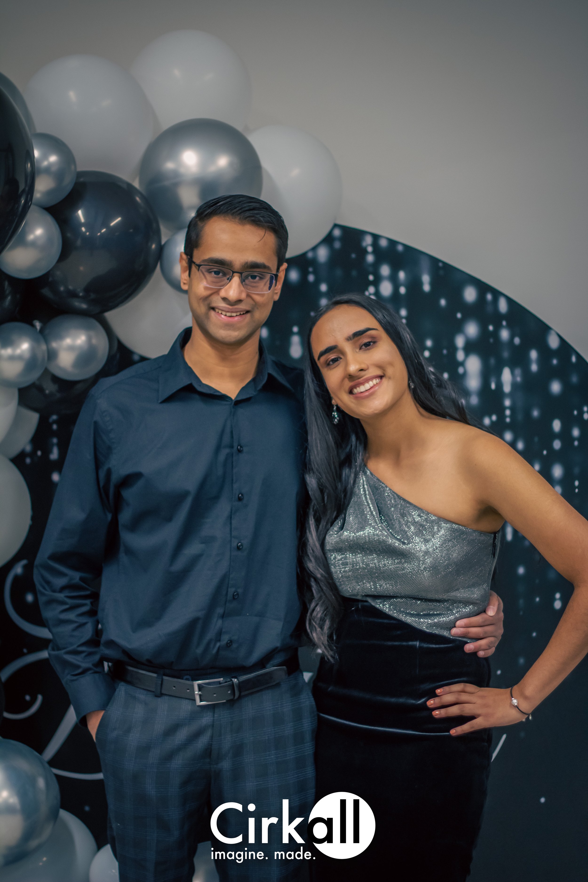 A young man and woman dressed formally, smiling at a celebration with a black and silver themed background. The man wears a navy blue shirt and plaid pants, and the woman wears a silver one-shoulder top and black skirt. There are balloons behind them