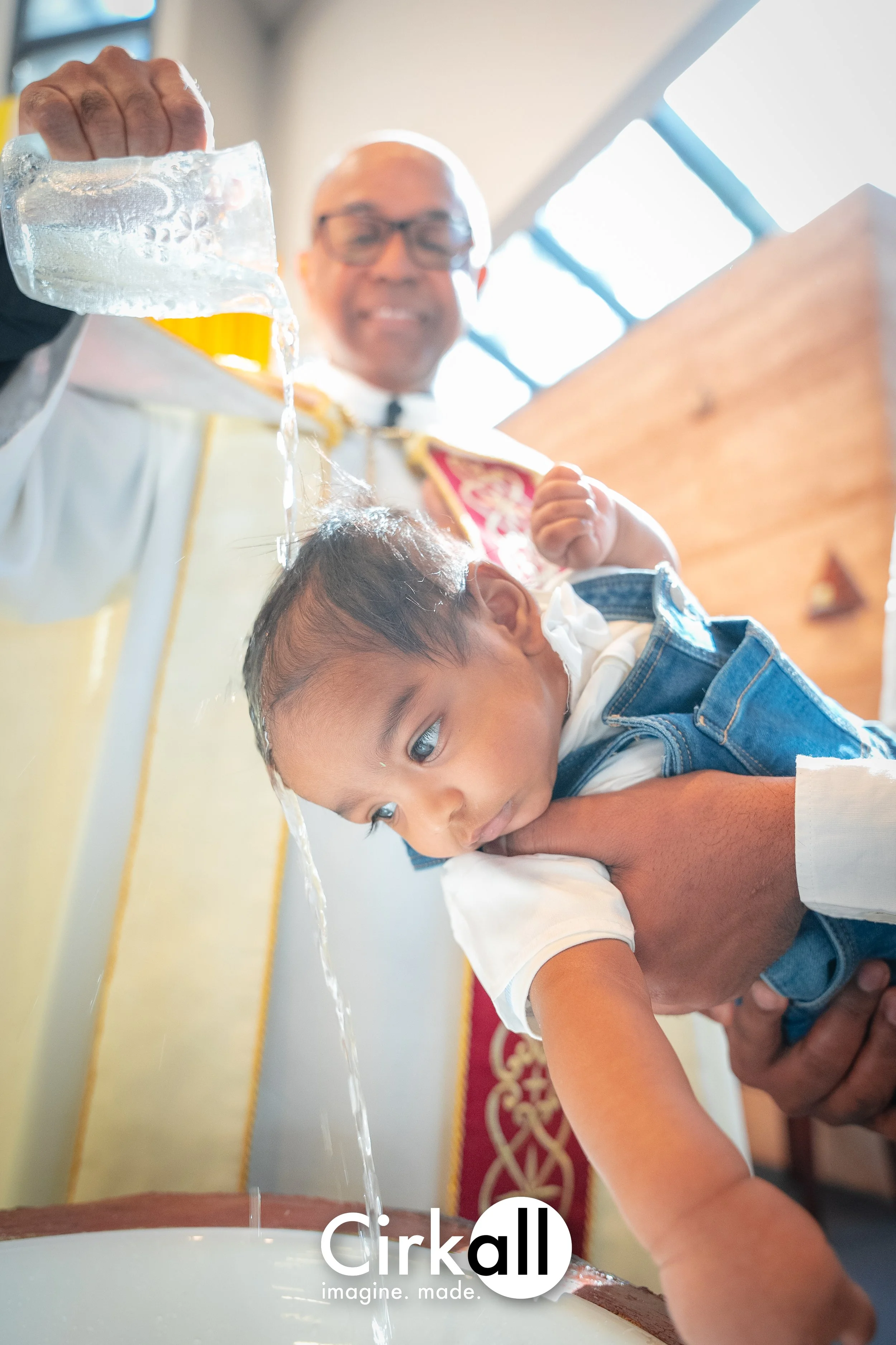 A baptismal ceremony with a priest pouring water over a child's head while holding the child, who looks curious. The priest is smiling and wearing ceremonial robes, with a wooden background and large windows in the background.