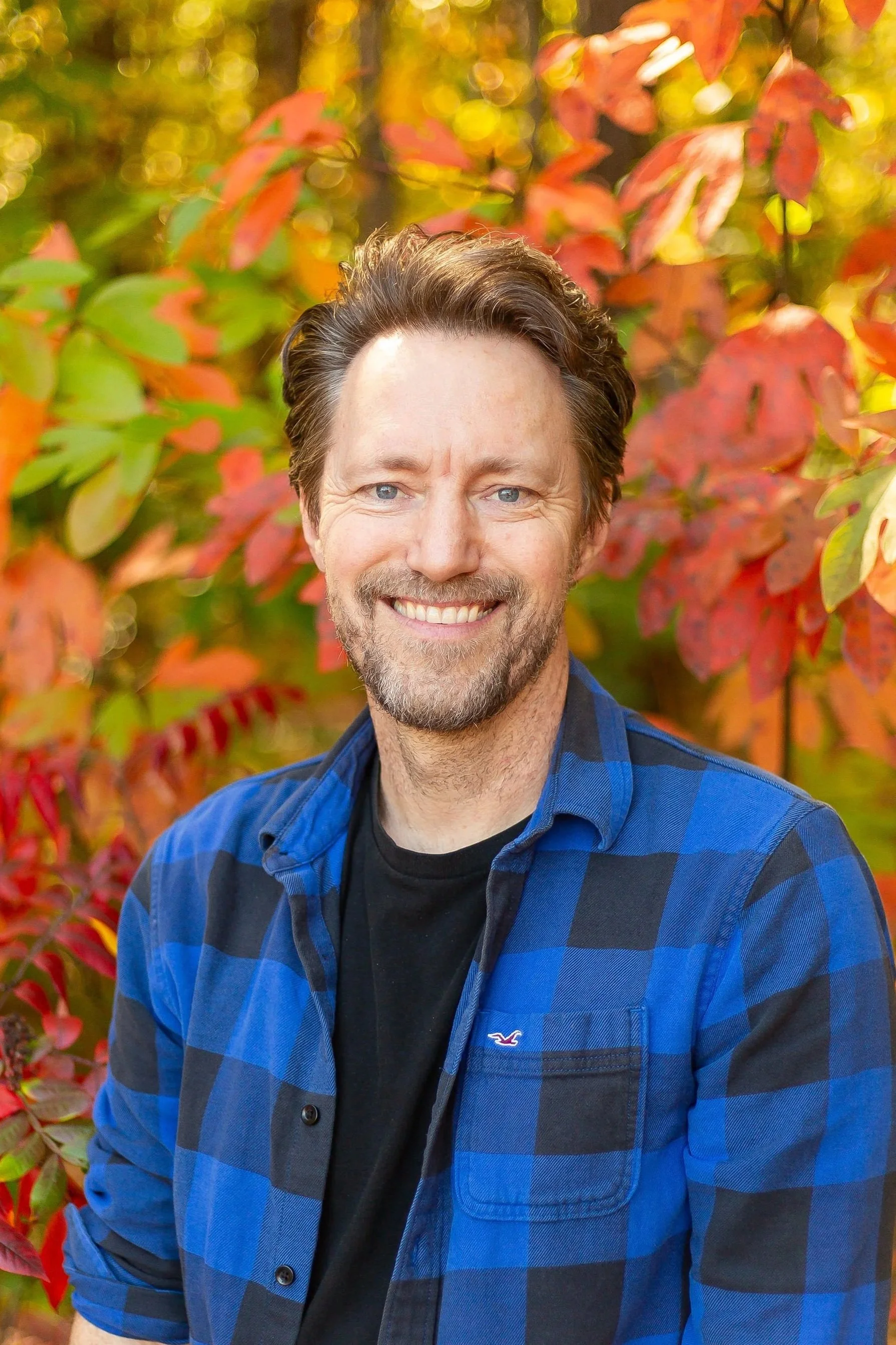 Smiling man with brown hair and beard, wearing a black t-shirt, sitting outdoors in a wooded area with green leaves.