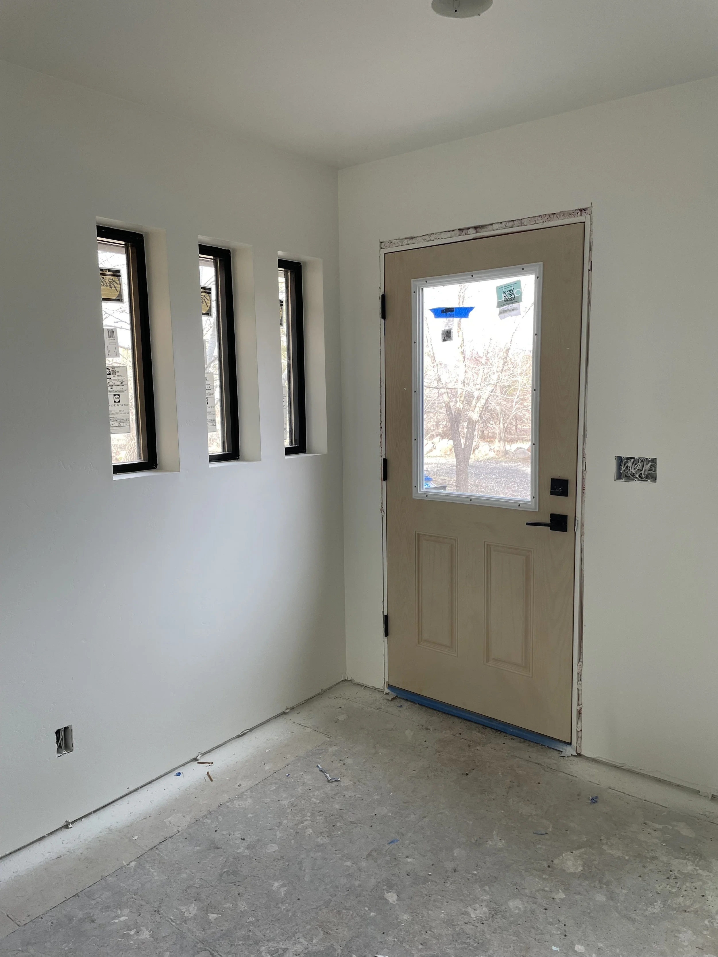 Interior view of a room under construction with a front door and three narrow windows to the left, unfinished floor and wall wiring.