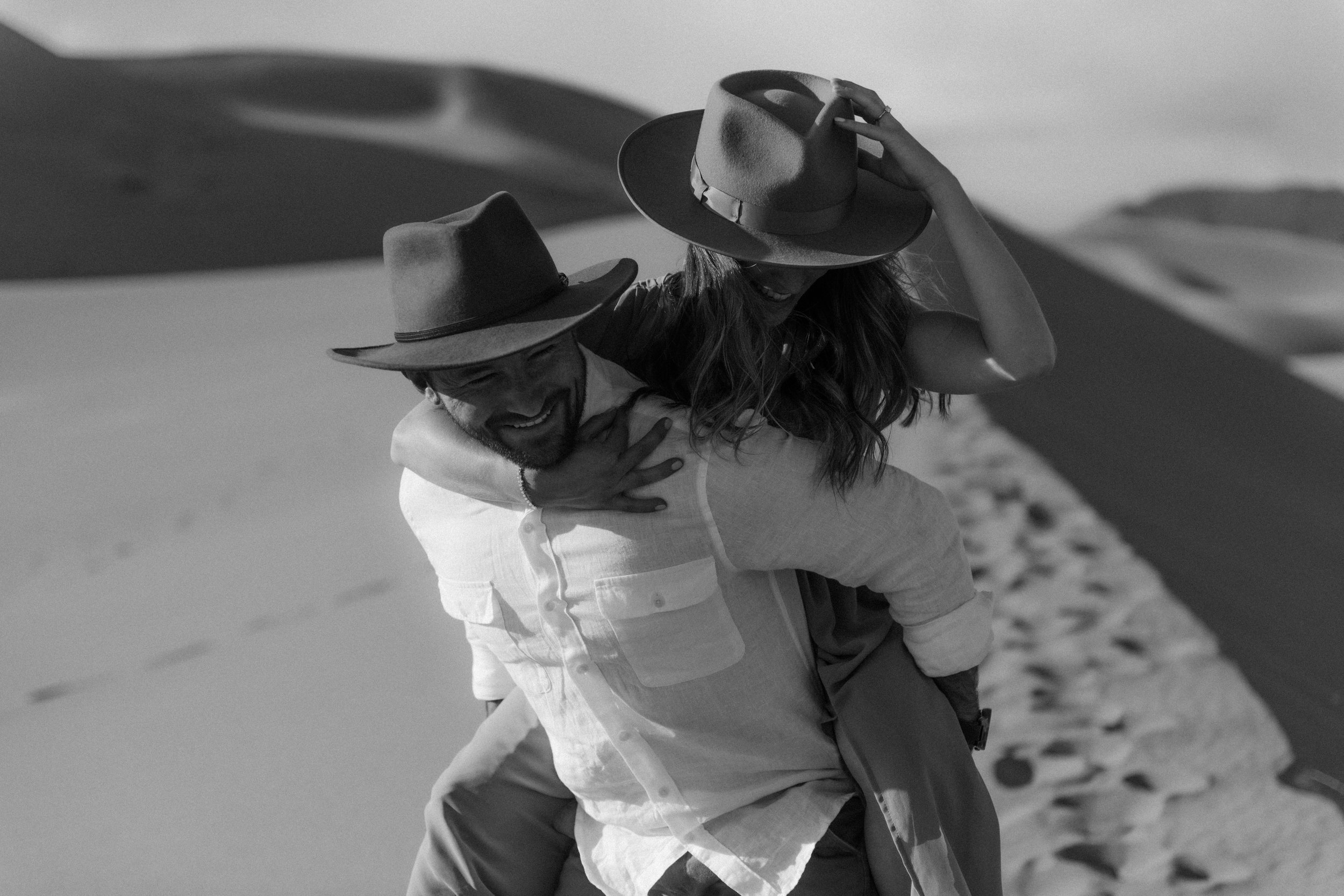 A black and white photo of a man and a woman at the beach, with the man giving the woman a piggyback ride. They are both wearing wide-brimmed hats, and the woman is adjusting her hat with her hand. The background shows sand dunes and the sky.