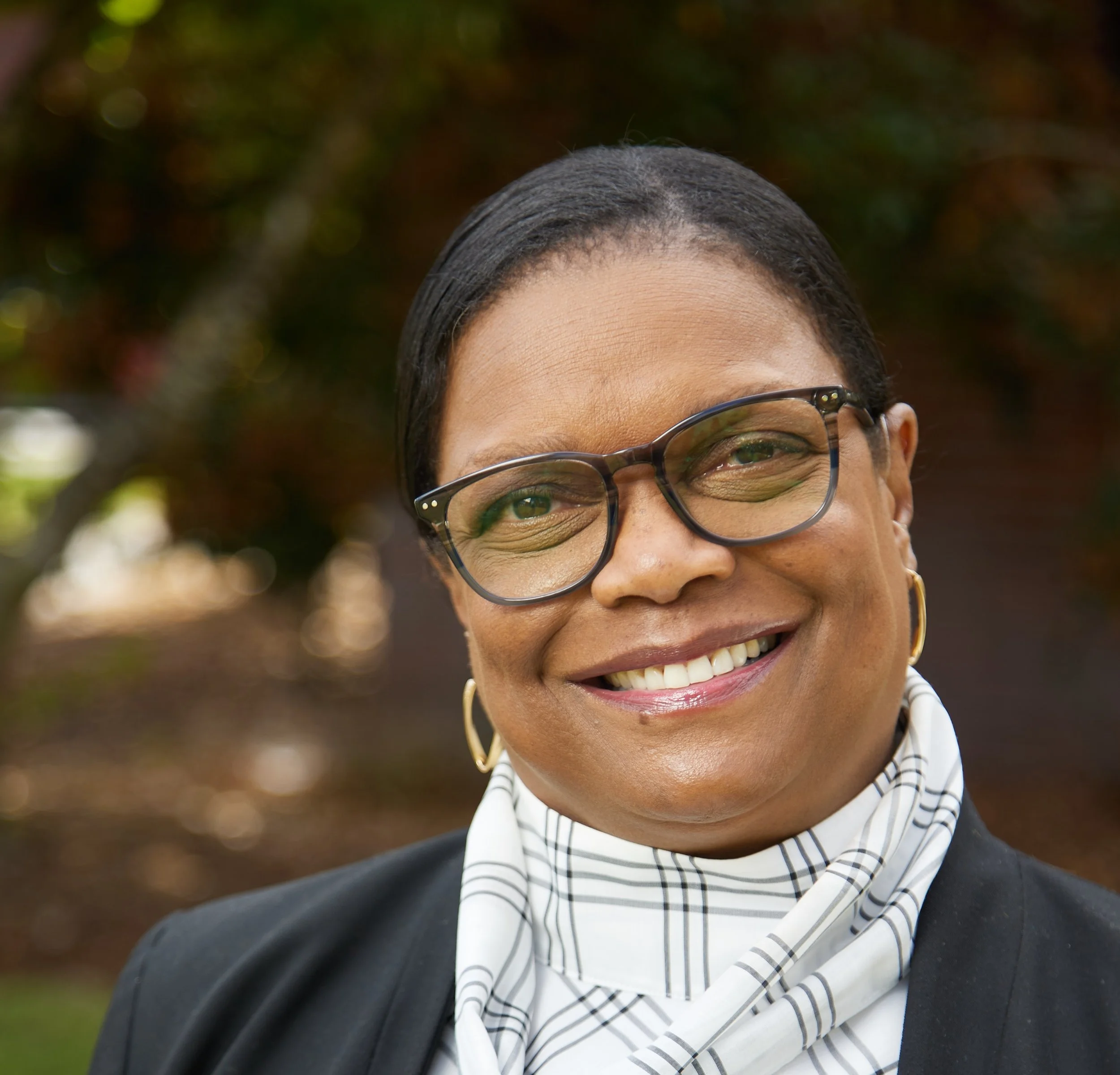 Close-up of an smiling woman with glasses, wearing a striped scarf and a black blazer, outdoors with blurred greenery background.