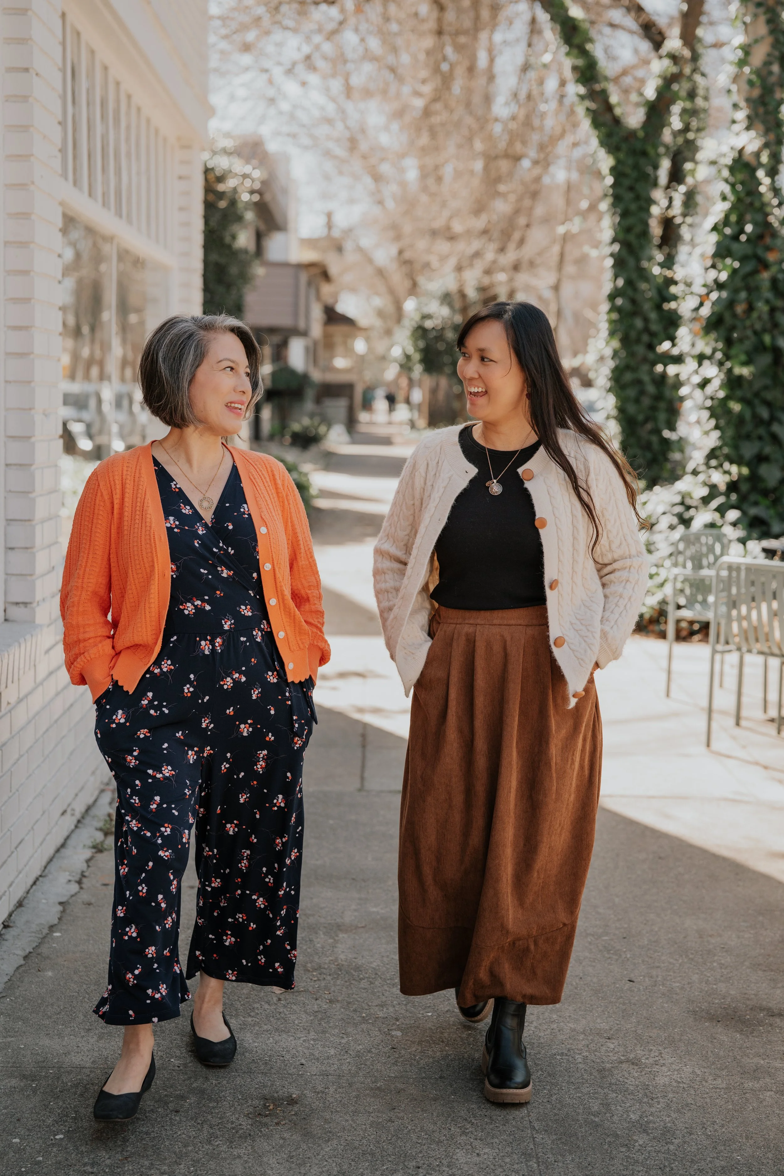 Two women walking and talking on a sidewalk. One woman has short gray hair, wearing an orange cardigan over a black floral dress, and black shoes. The other woman has long dark hair, wearing a white knit jacket, black top, brown wide-leg pants, and black boots. The background shows trees and residential buildings.
