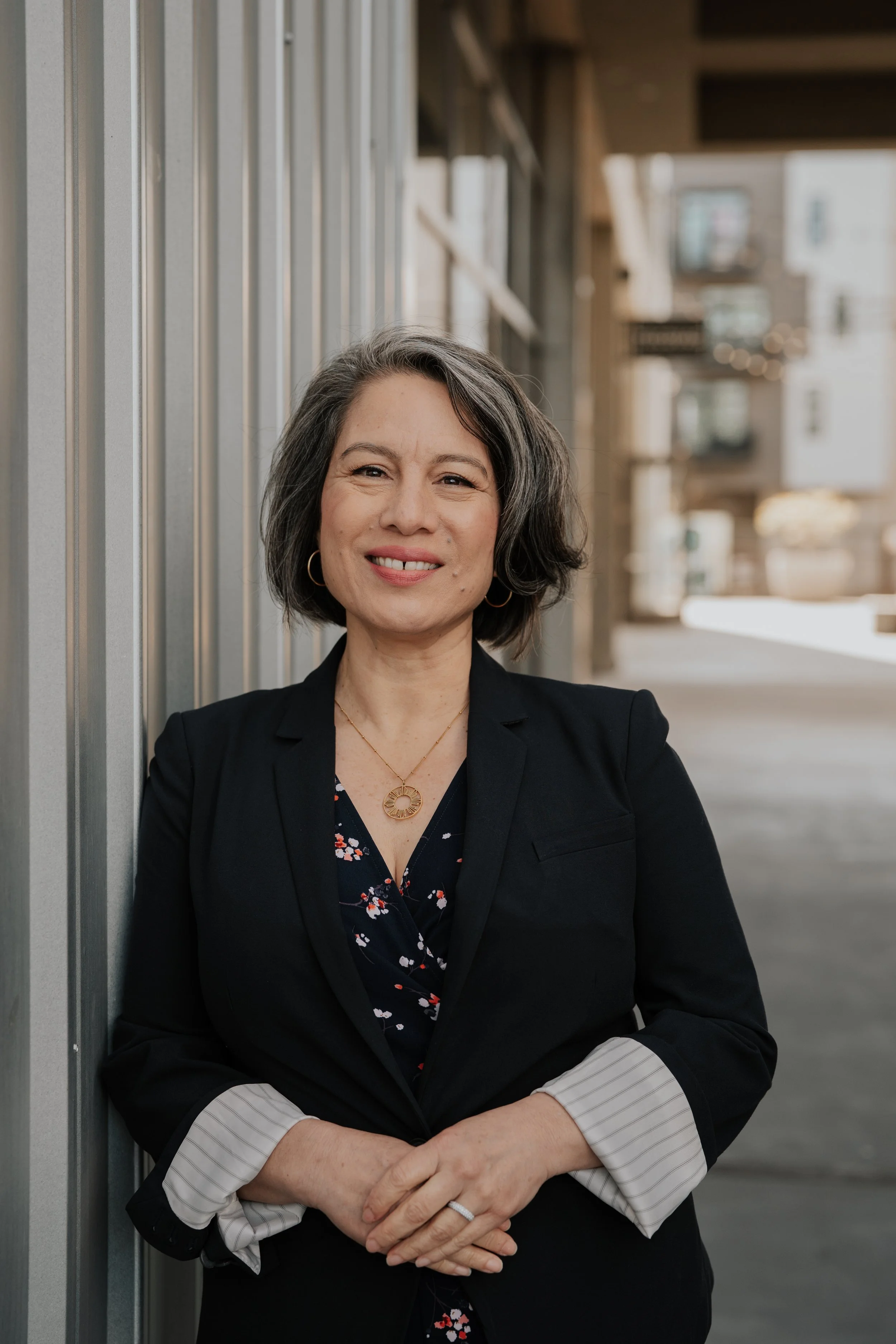 A woman with shoulder-length gray hair, wearing a black blazer over a floral blouse, smiles and stands with arms crossed in an urban outdoor setting with buildings in the background.
