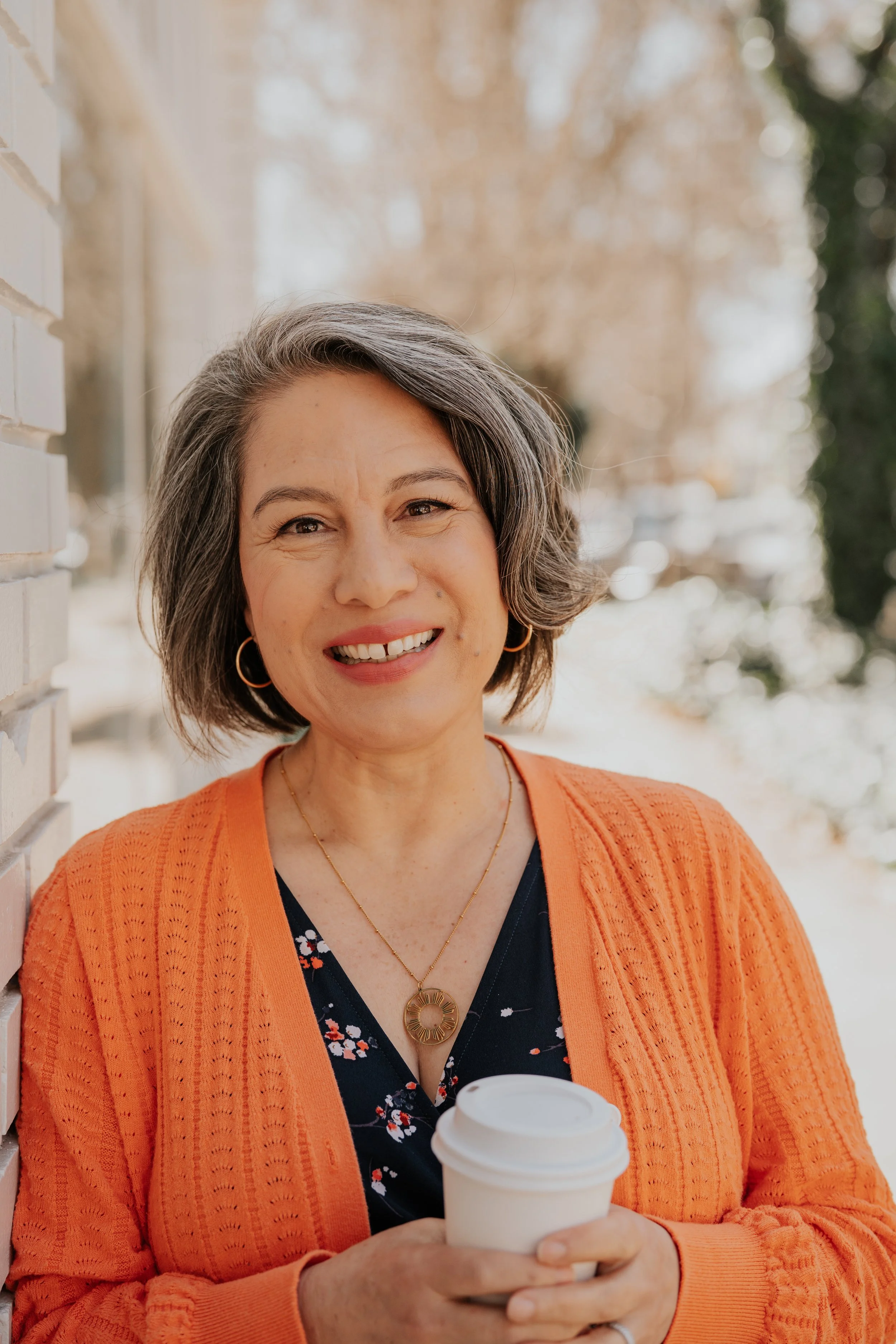 A woman with short gray hair wearing an orange cardigan and a navy floral blouse holding a white disposable coffee cup outdoors on a sunny day.
