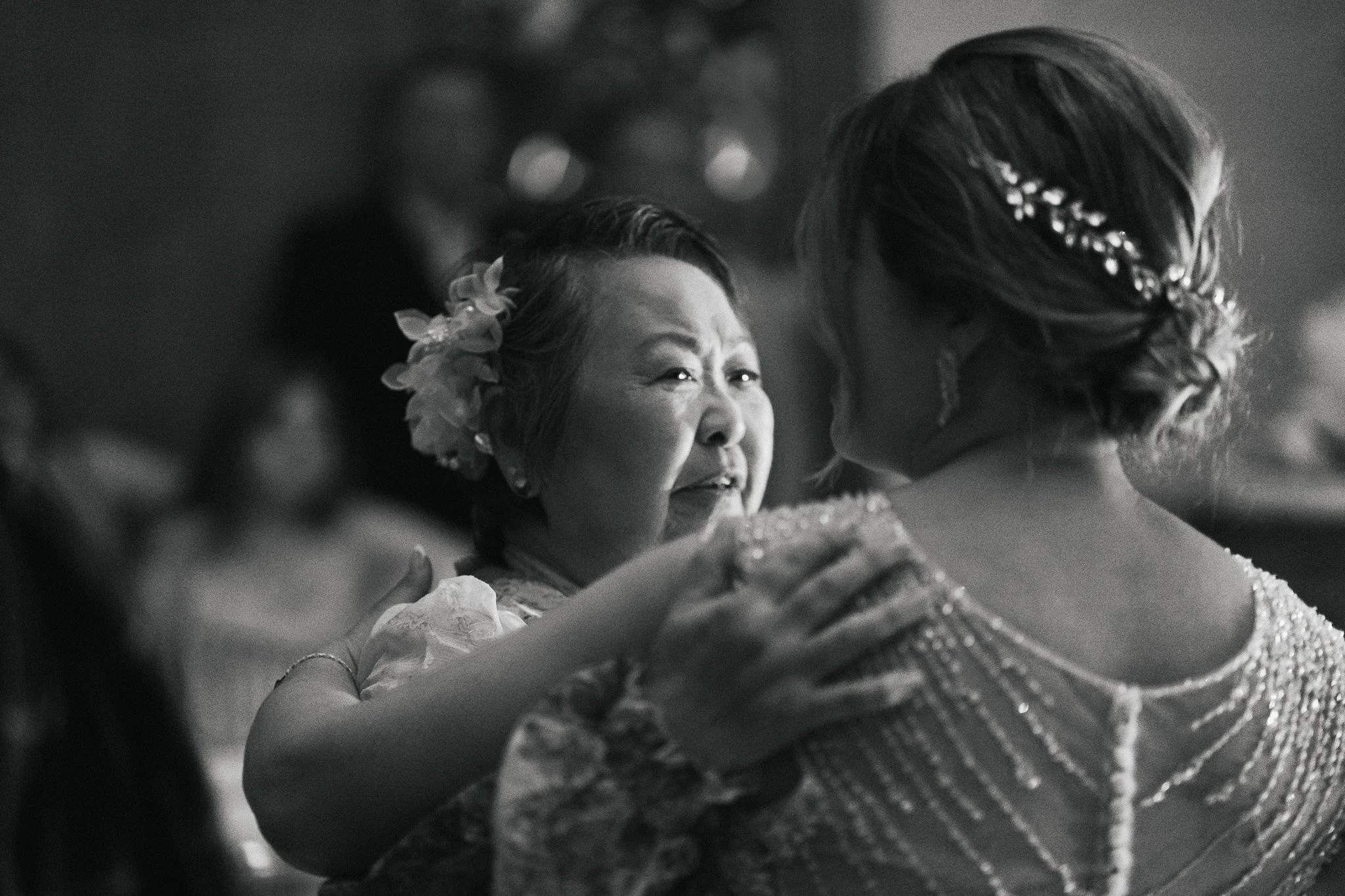 A black and white photo capturing a close-up moment between two women, one older with flower earrings and the other younger with hair decorated with a pearl hairpiece, possibly sharing a kiss or embracing.