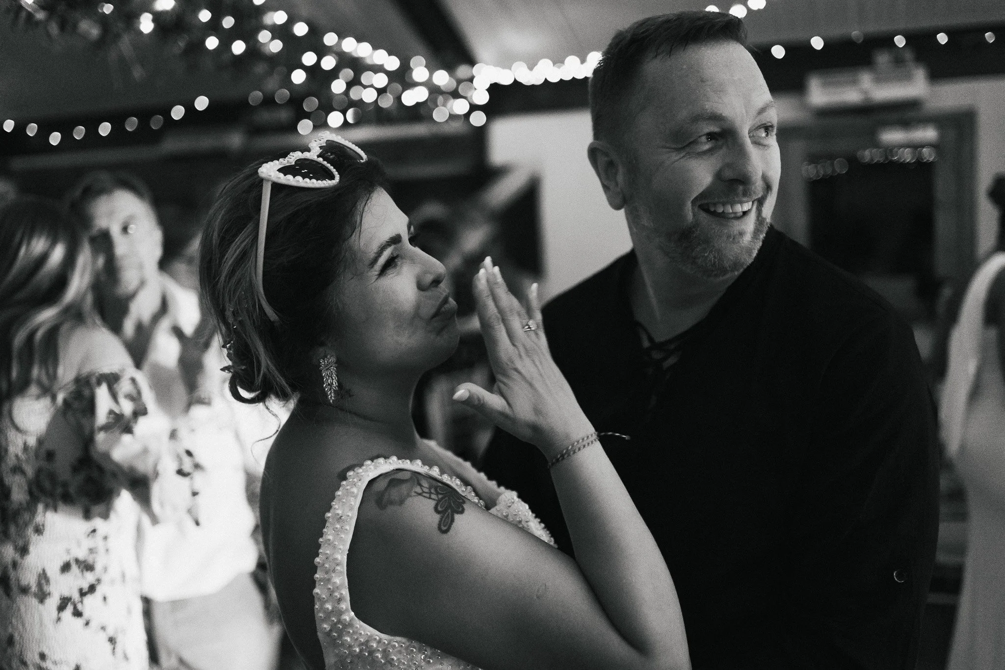 A black and white photo of a group of people at an event, with a focus on a smiling woman with dark hair, tattoos on her arm, and wearing heart-shaped sunglasses on her head. A man with short hair and a beard is next to her, smiling and looking to th