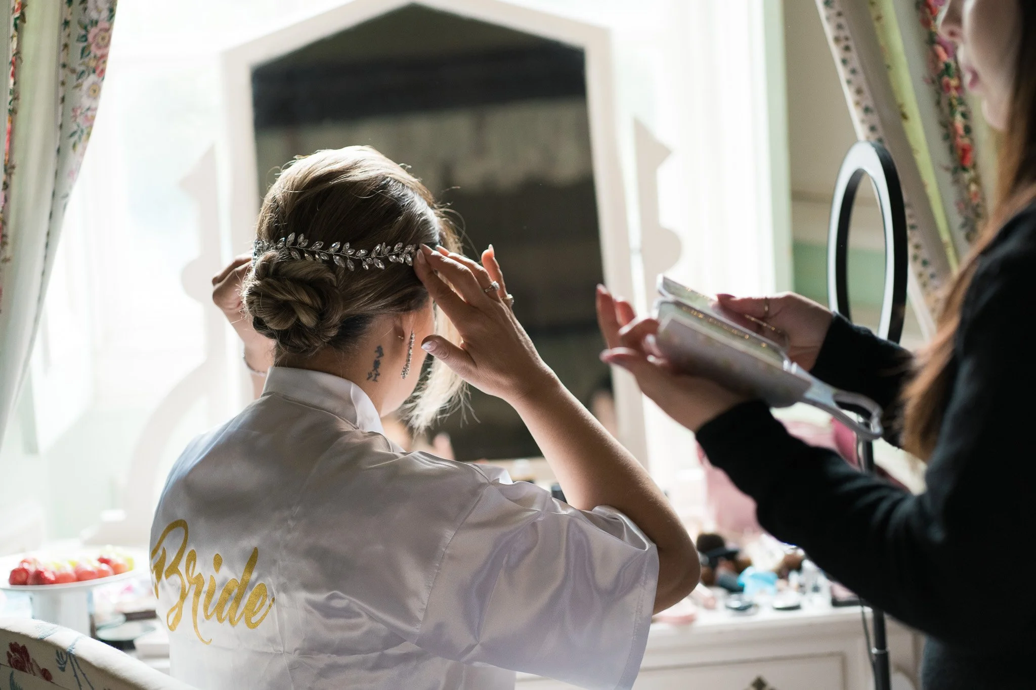 Bride getting ready, wearing a 'Bridal' gown and a jeweled headpiece, as a woman adjusts her hair in front of a mirror.