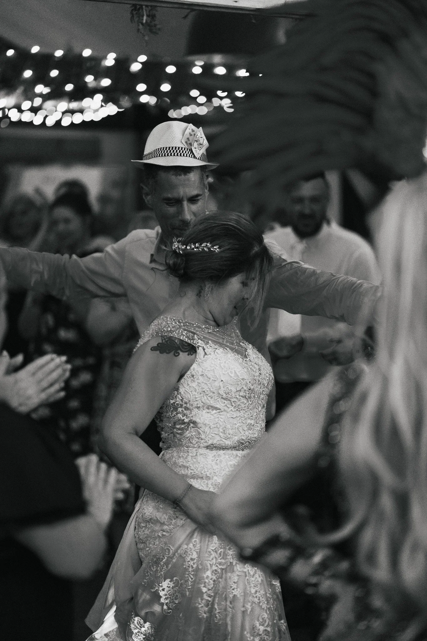 A woman in a wedding dress with lace details is dancing with her arms outstretched, surrounded by a group of people, some of whom are clapping and smiling. The scene is decorated with string lights hanging from the ceiling.